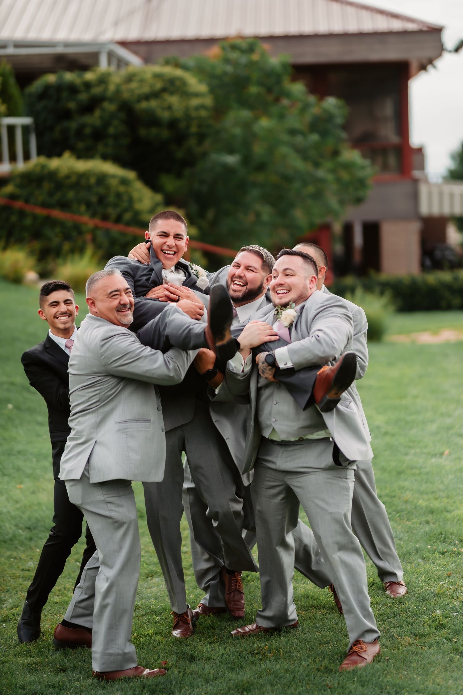 Wedding party members in gray suits create a fun group pose while laughing on green grass.