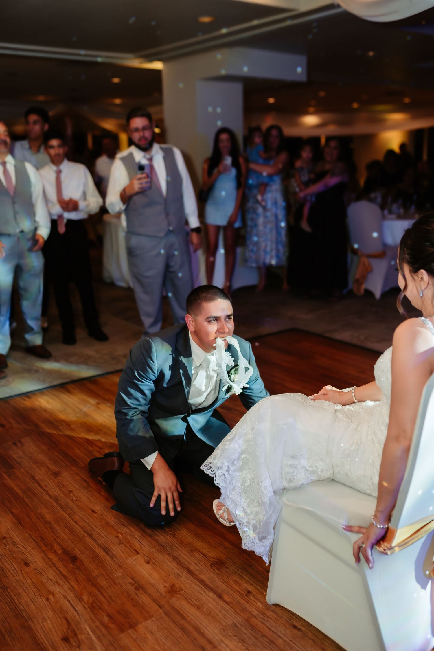A wedding reception scene showing traditional garter removal ritual on a wooden dance floor.