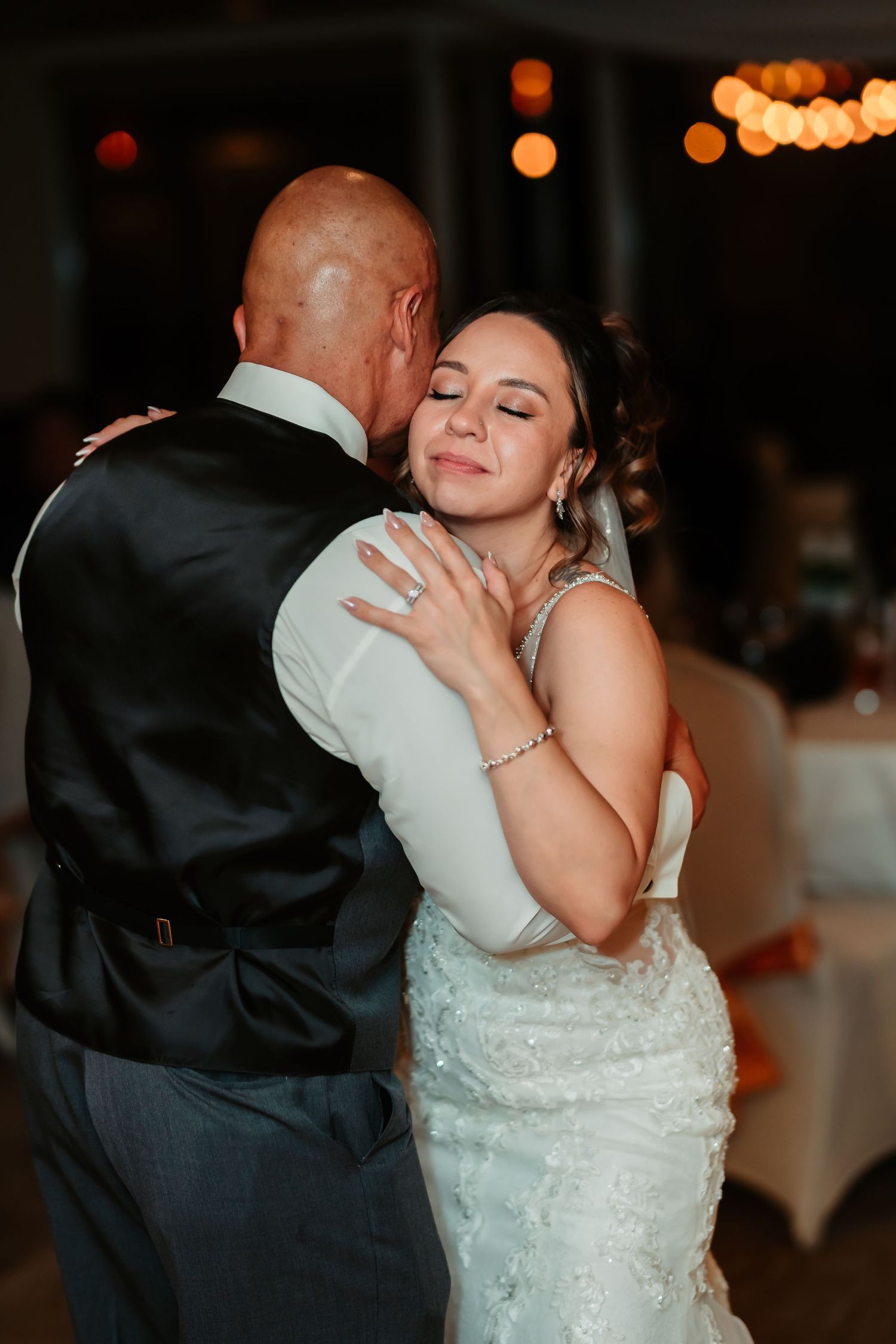 Two people share a romantic dance at a wedding reception with soft bokeh lights in the background.