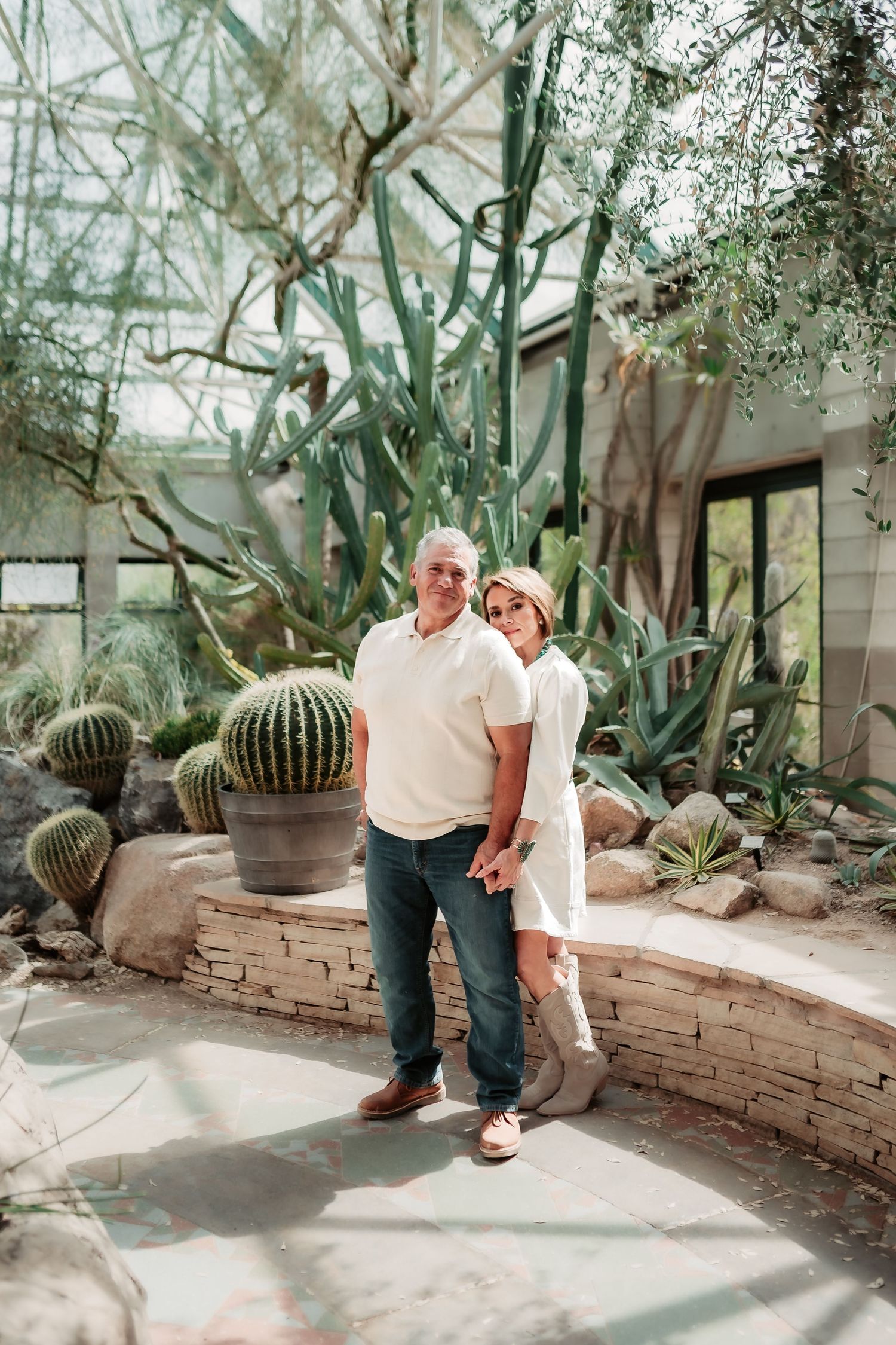 A couple shares a tender moment in a greenhouse surrounded by desert plants and stone walls.