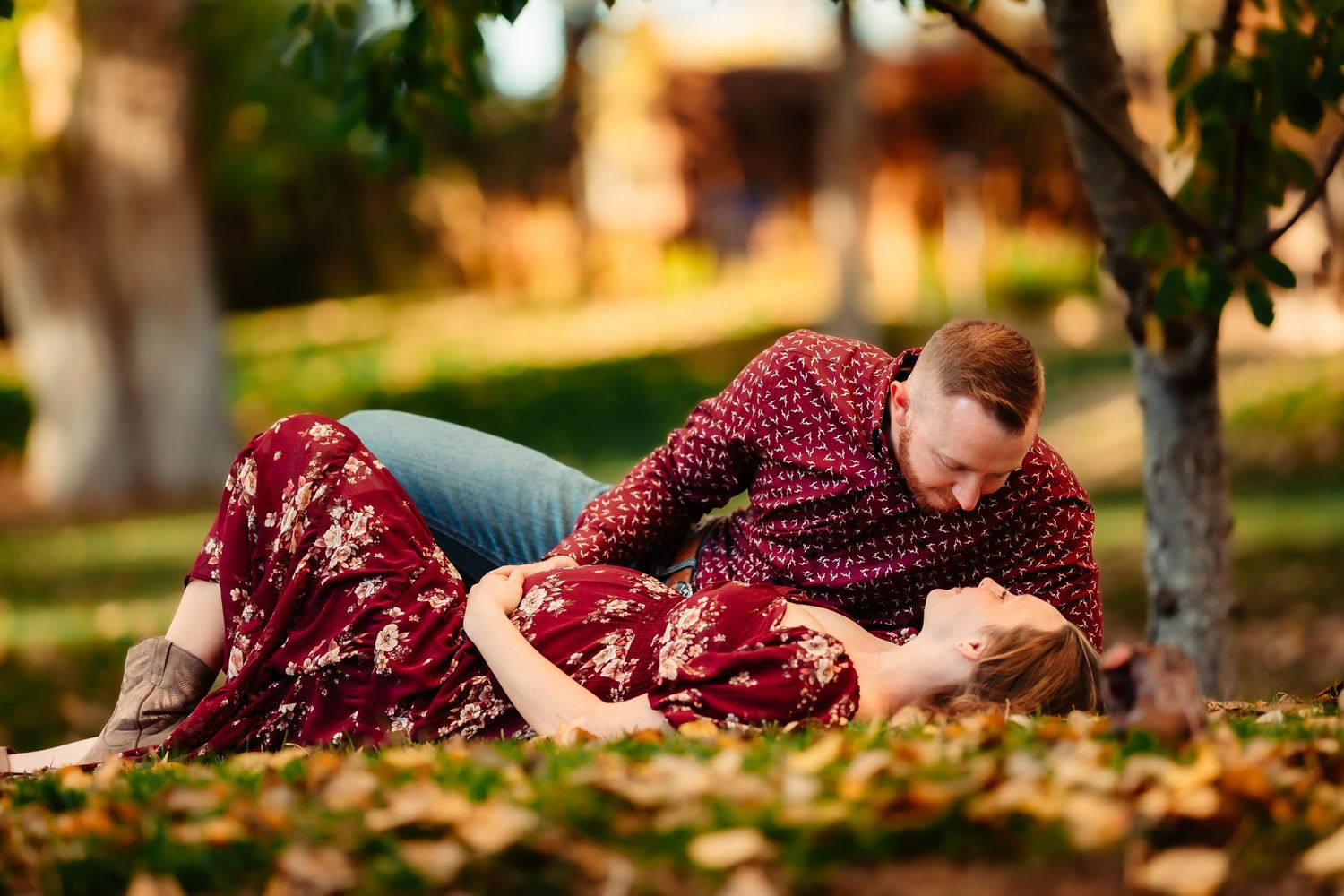 A person in a burgundy dress lies gracefully among scattered autumn leaves during a golden hour photoshoot.