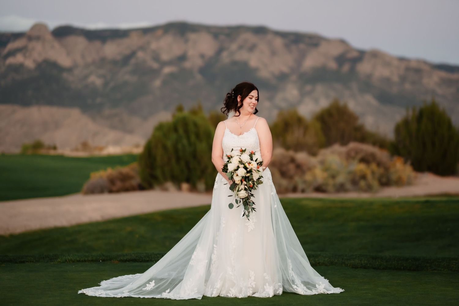 A bride in a flowing white gown holds a bouquet while standing on a golf course with mountain views at sunset.