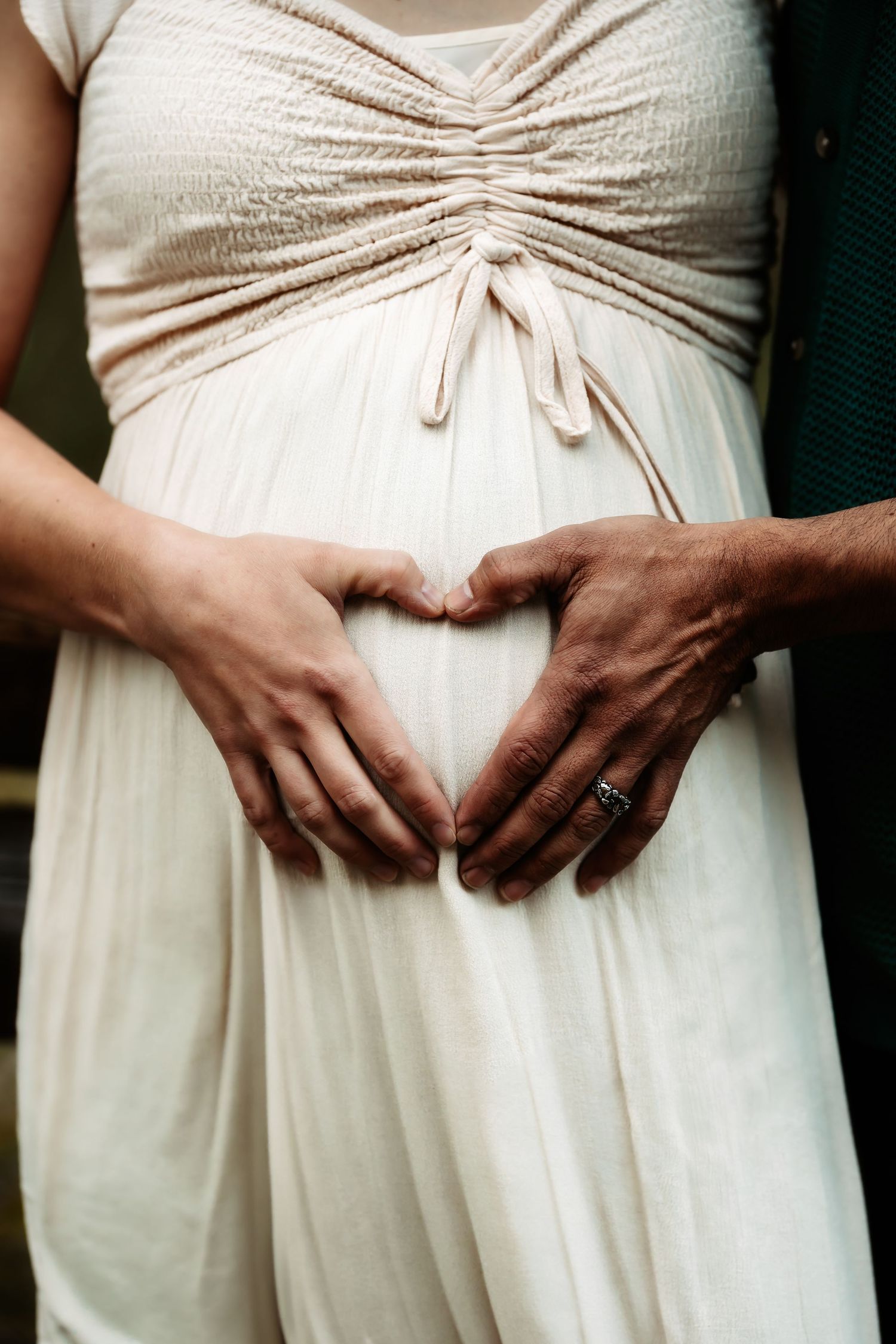 Two hands form a heart shape on a pregnant belly wearing a ruched white maternity dress.