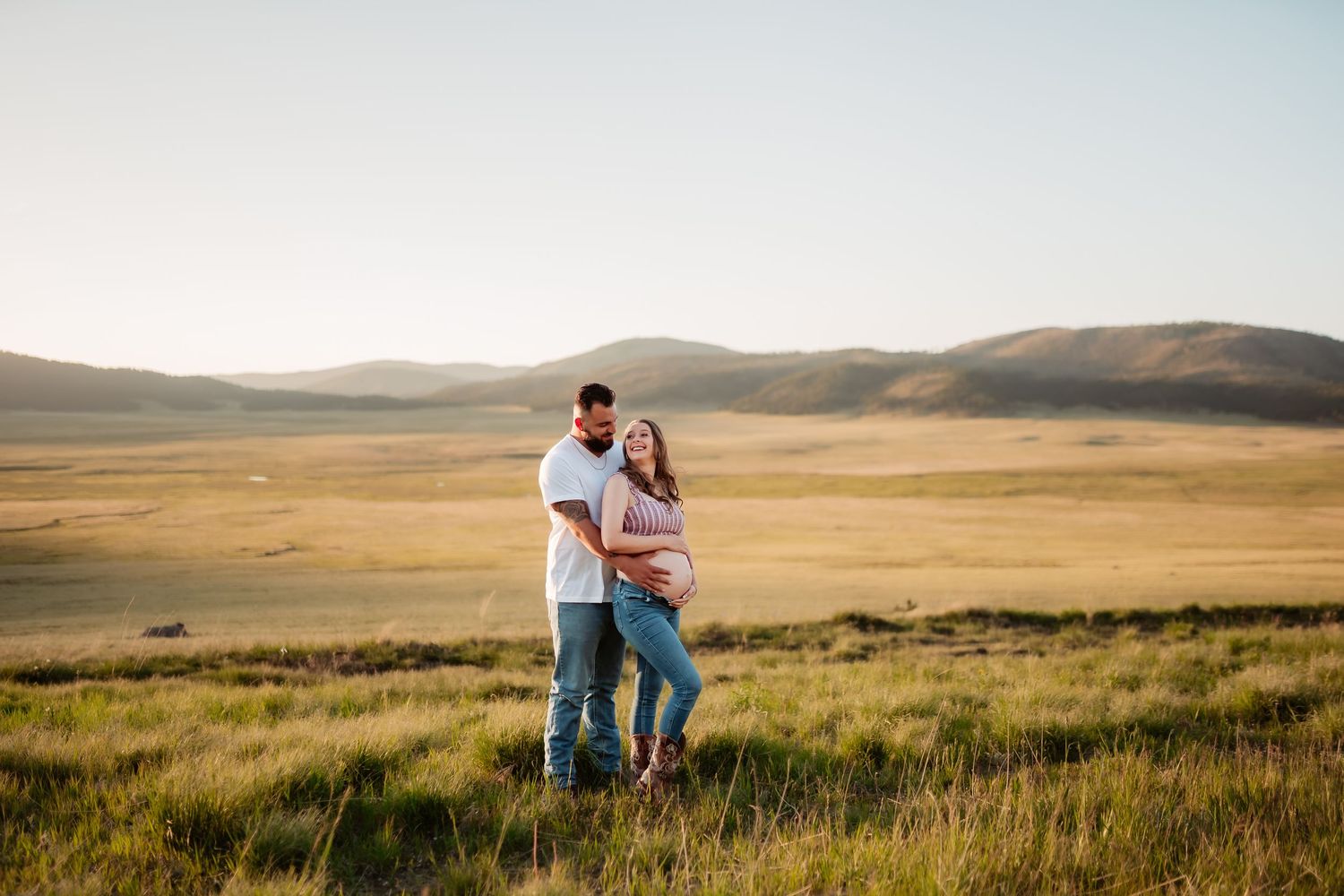 A couple embraces in a golden field at sunset with rolling hills and mountains in the distance.