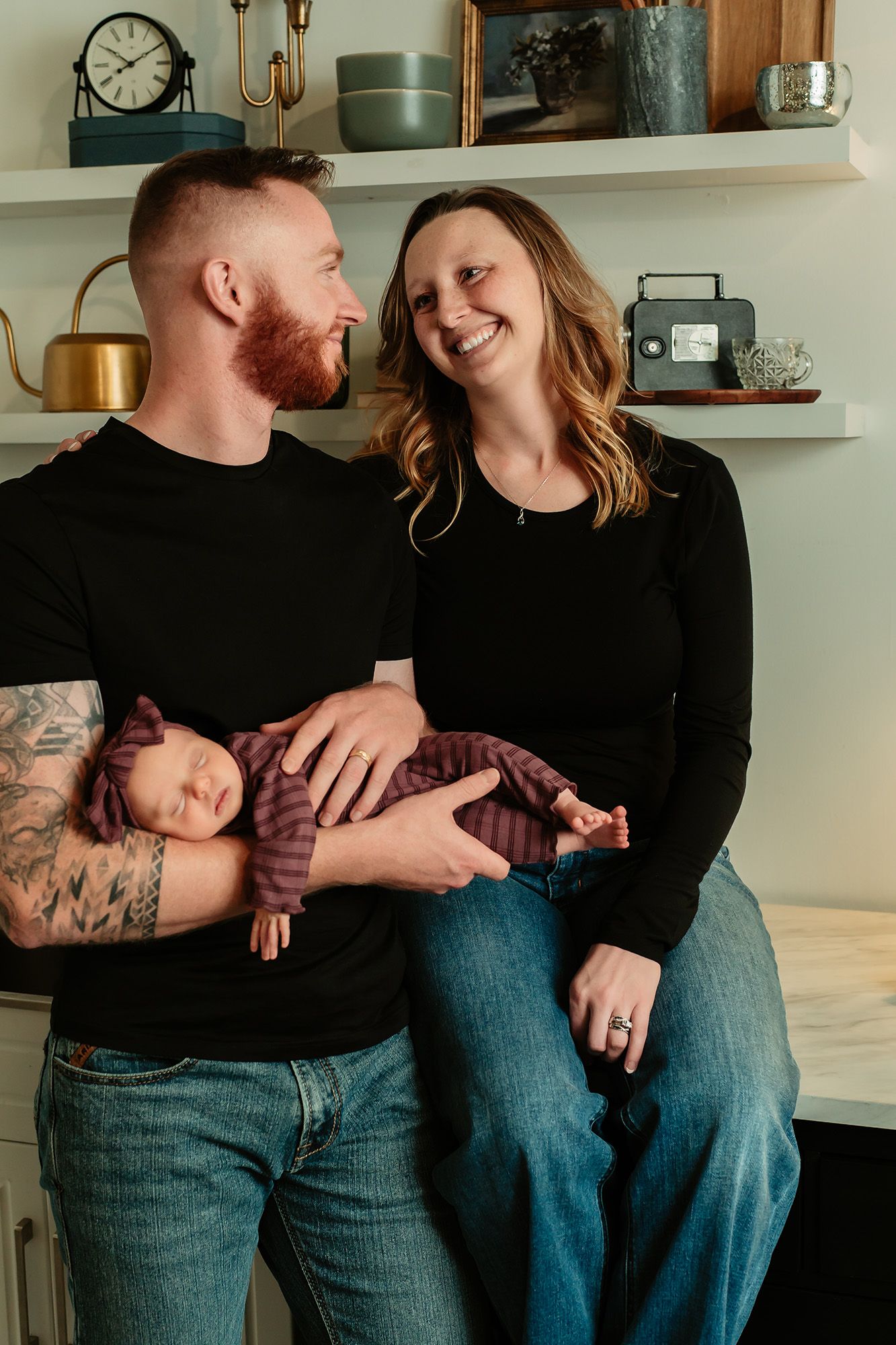A smiling couple in black tops and jeans holds their sleeping newborn wrapped in purple in a cozy home setting.