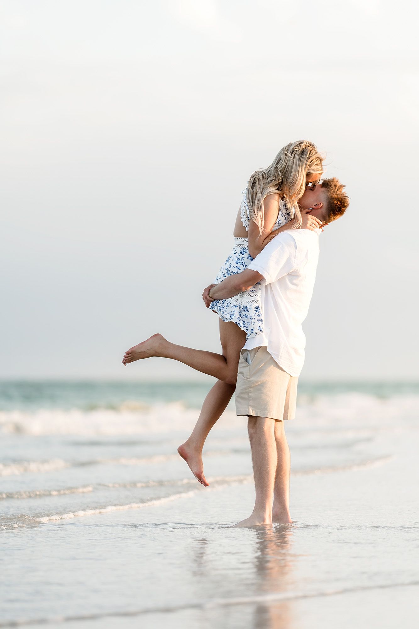 Romantic couple embracing and kissing on a beach at sunset, man lifting woman in white dress near ocean waves.