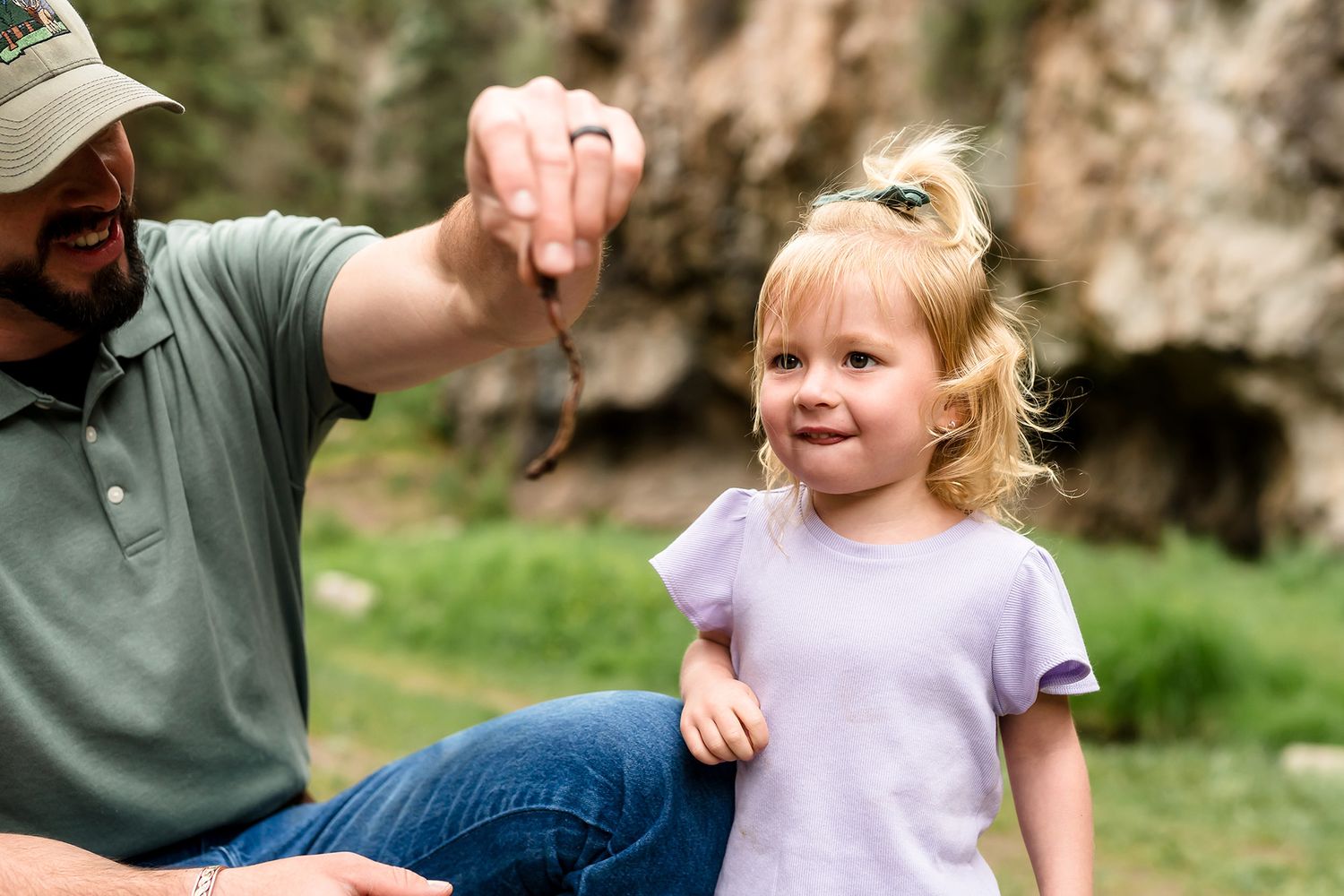 A father holds a small snake near his curious toddler daughter outdoors in a rocky nature setting.