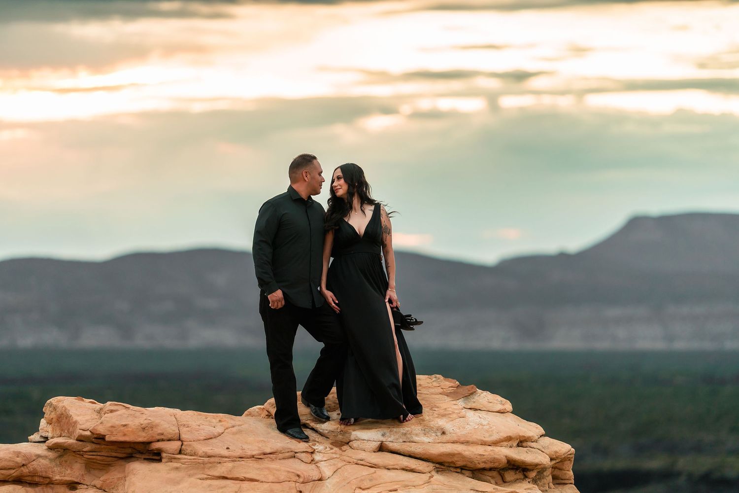A couple in matching black outfits share a romantic moment on desert sandstone rocks at golden hour.