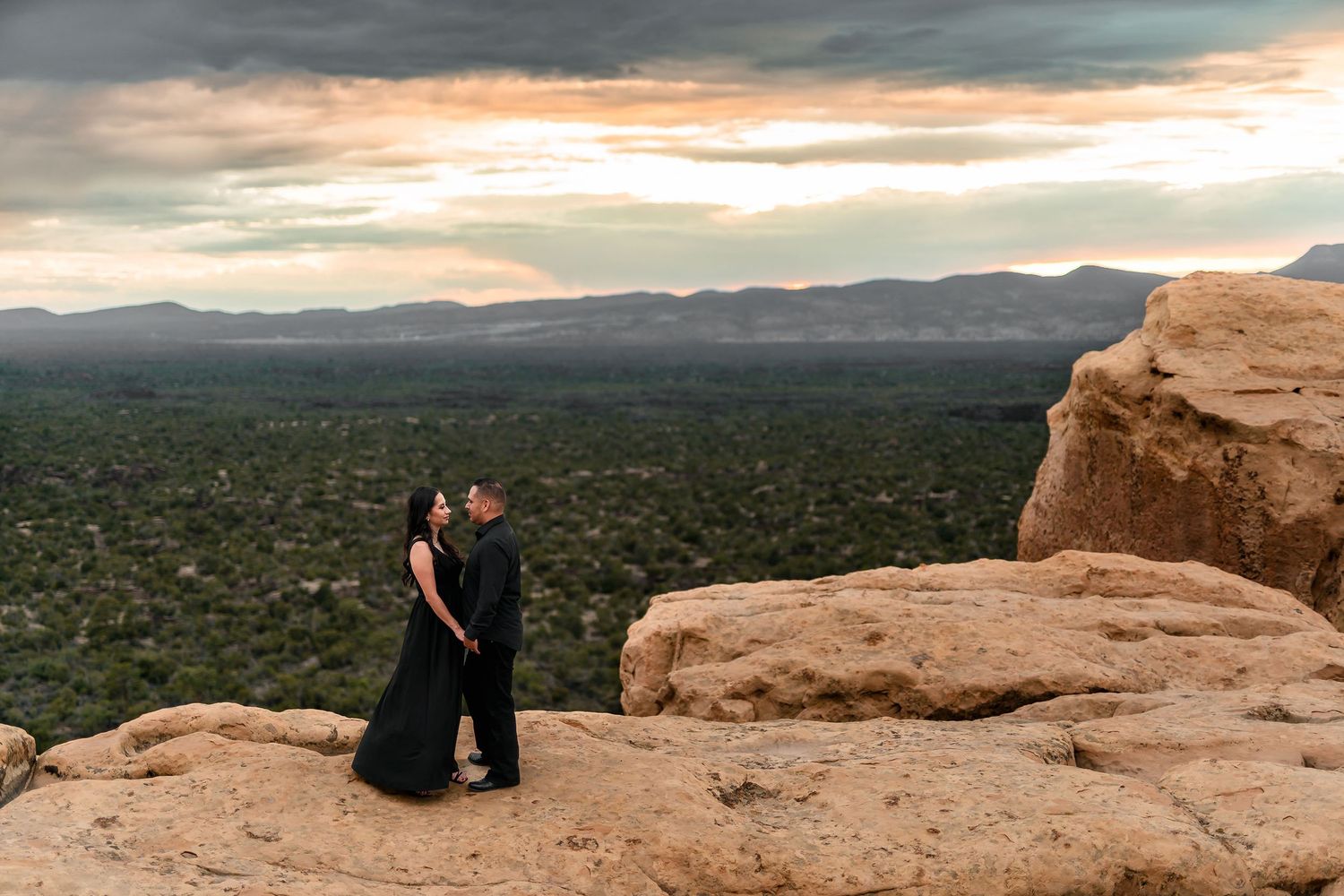 Couple in black attire embracing on rocky desert overlook at sunset with mountain valley views.
