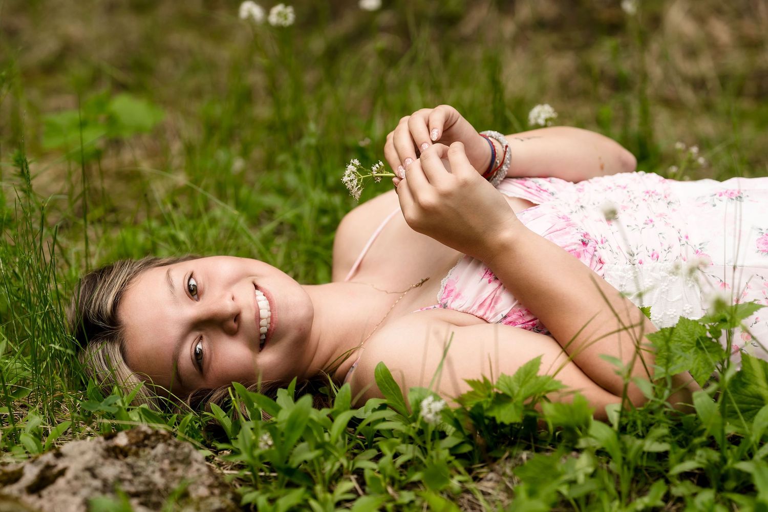 Smiling young woman in pink dress lies in green clover field holding a small white flower.