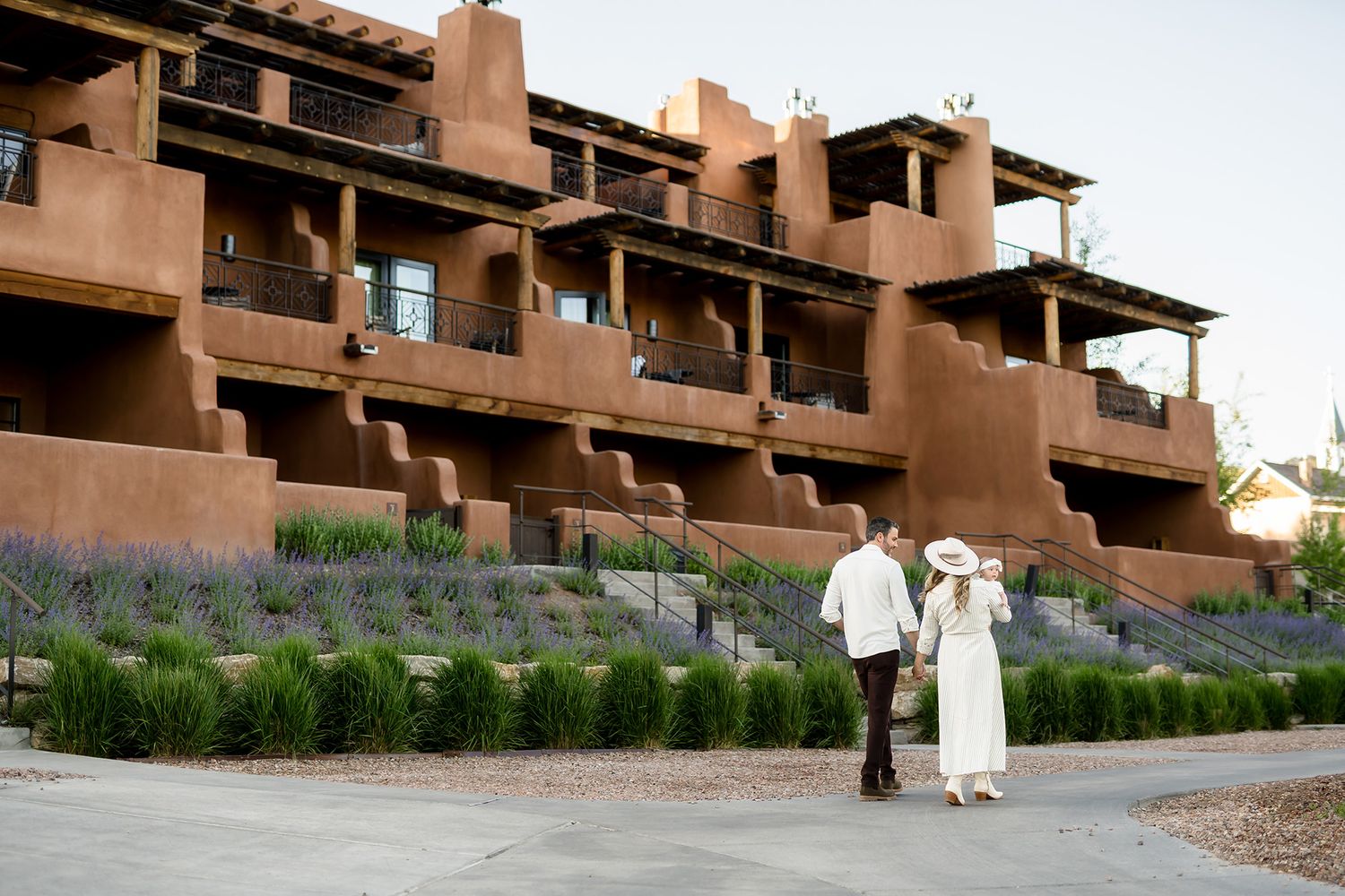 Couple holding hands walking toward a Southwestern adobe resort with tiered balconies and lavender gardens.