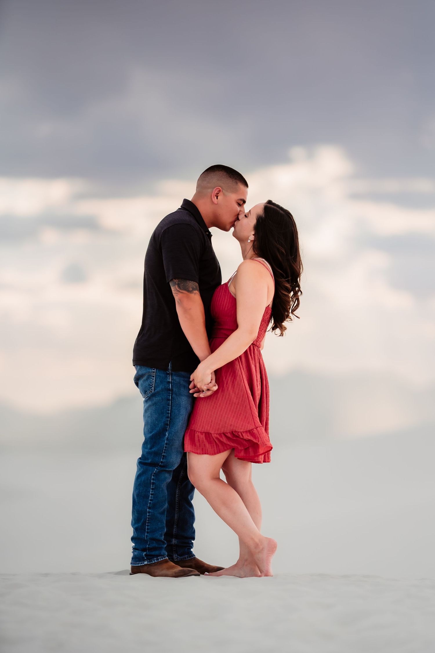 A couple embraces tenderly on a white sandy beach at sunset, with her in a red dress and him in a black shirt.