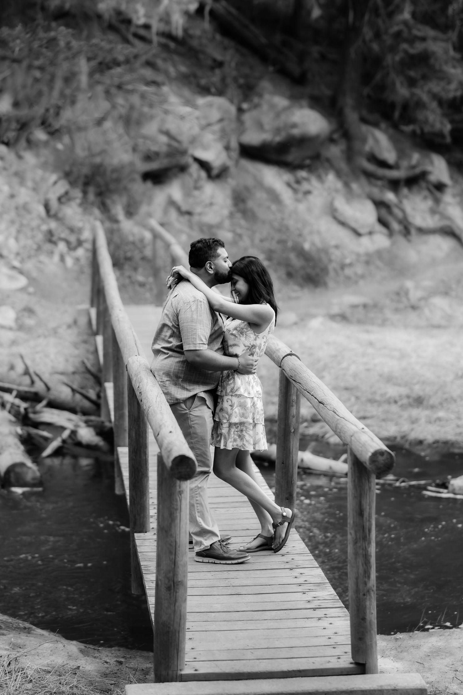 A tender moment between a couple on a wooden footbridge in a scenic outdoor setting.