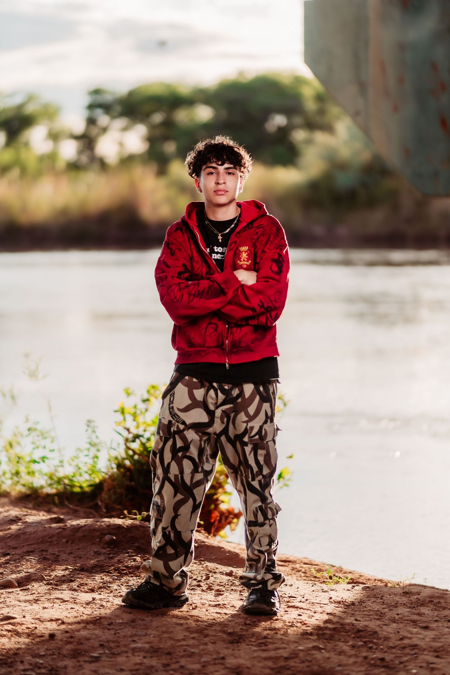 A person in a red hoodie and camo pants stands confidently by a riverbank in warm sunset lighting.