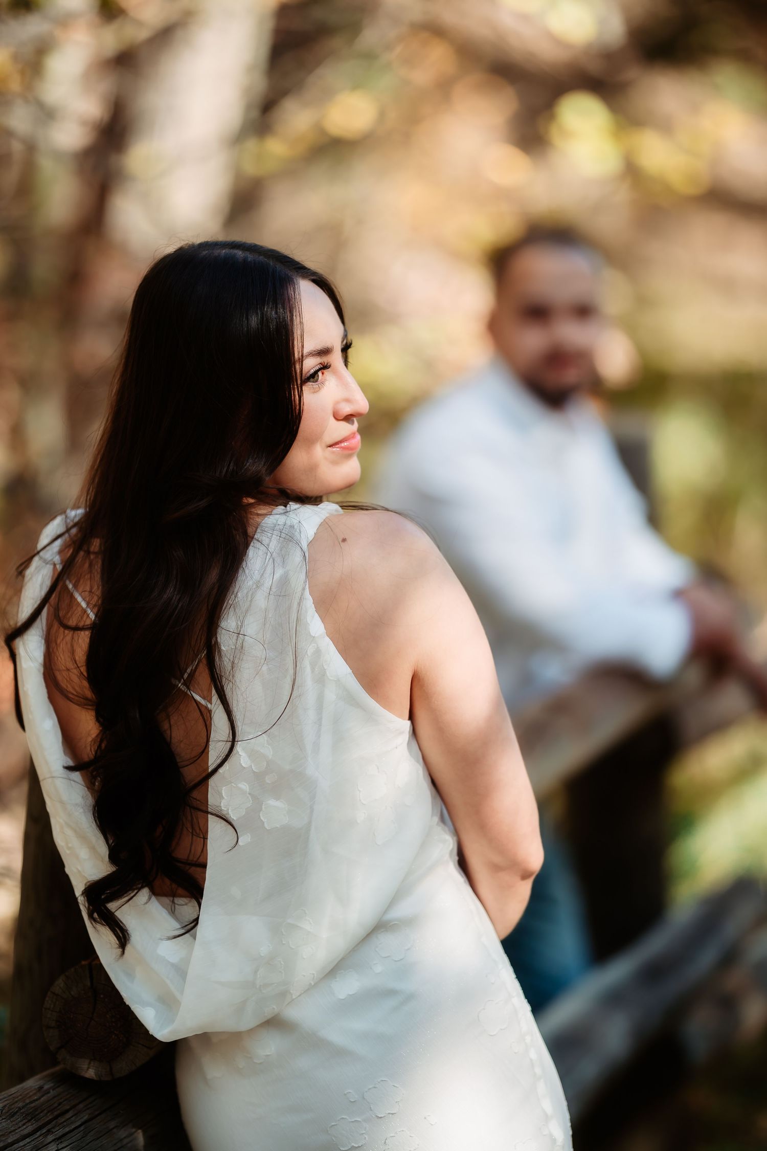 Woman in a white dress smiles and twirls outdoors in a natural setting with soft bokeh background.
