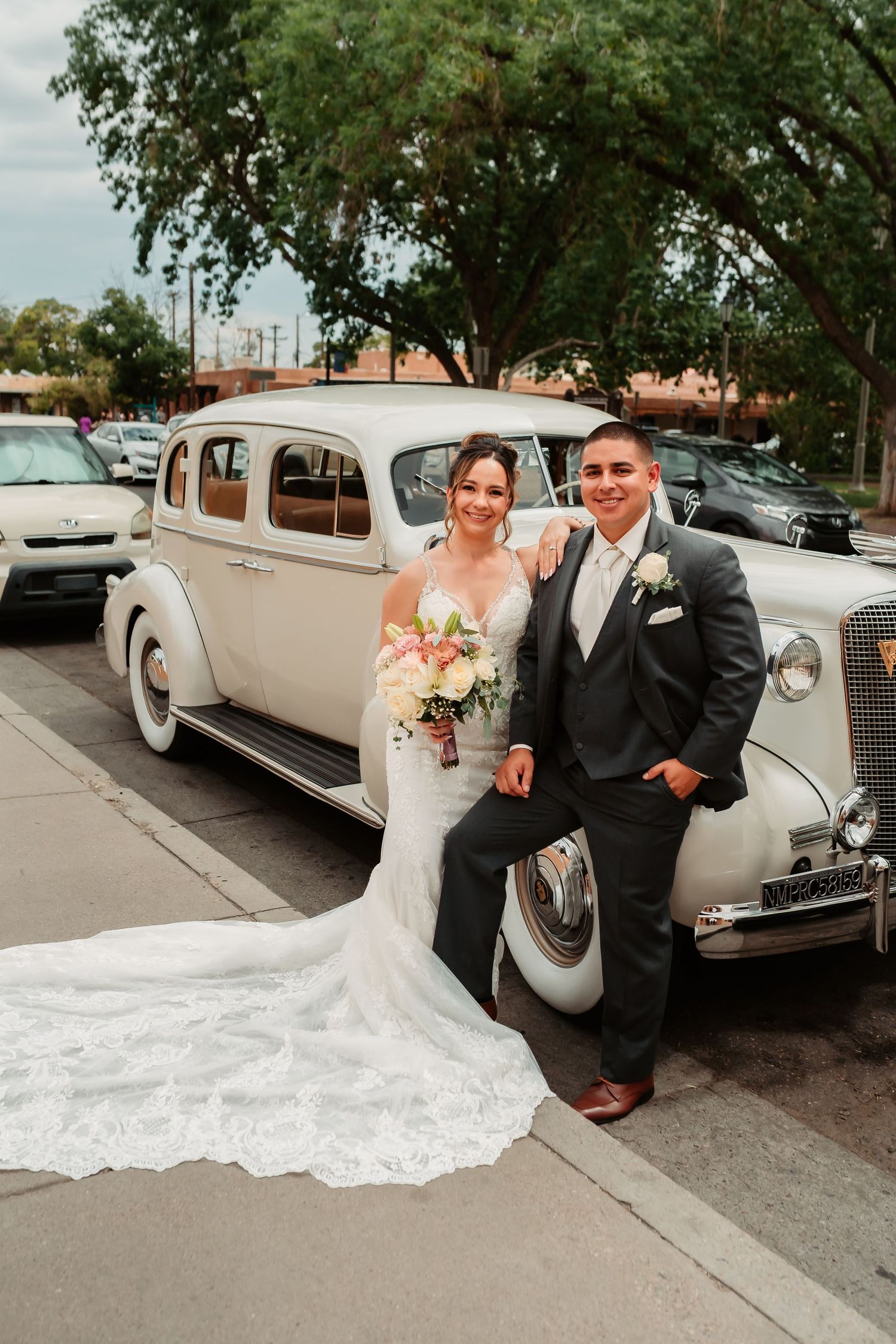 Newlyweds pose with a classic vintage cream-colored car on their wedding day.