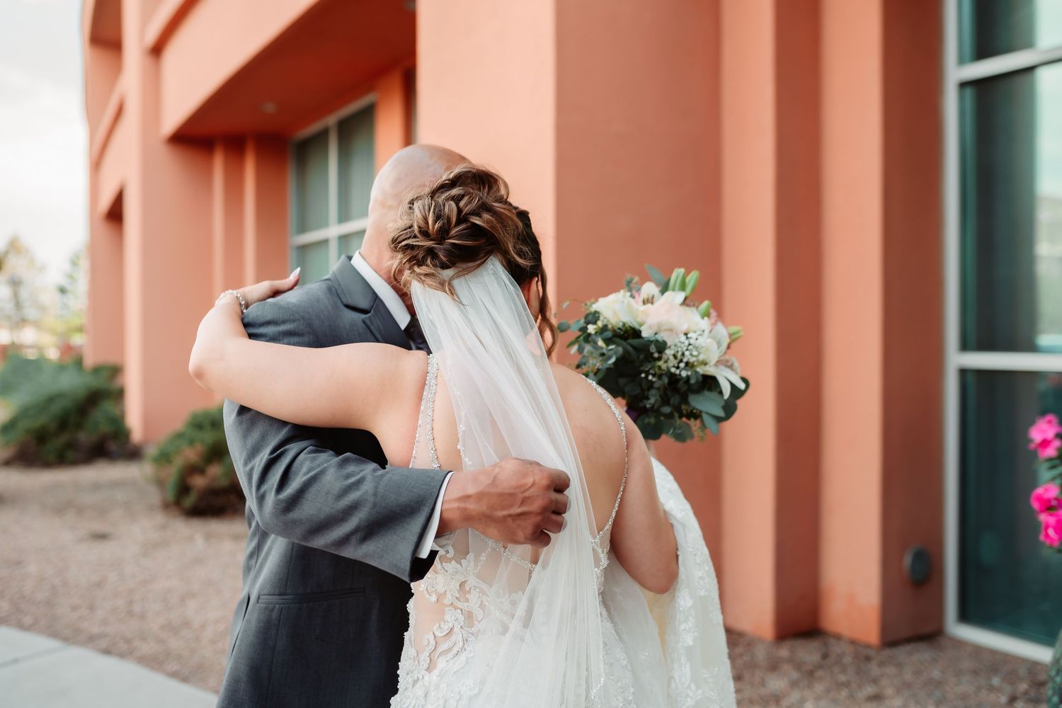 A couple in gray and white wedding attire embraces on a stone paved walkway by a coral colored building.