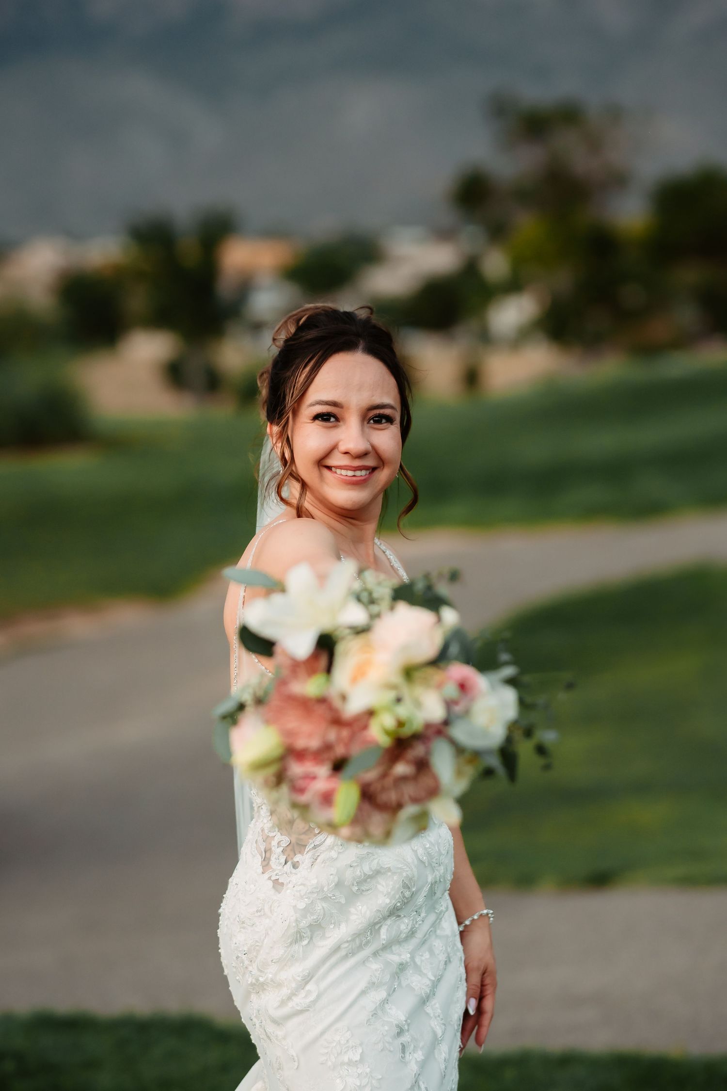Elegant bride in lace dress poses with beautiful bouquet of blush and ivory flowers outdoors.