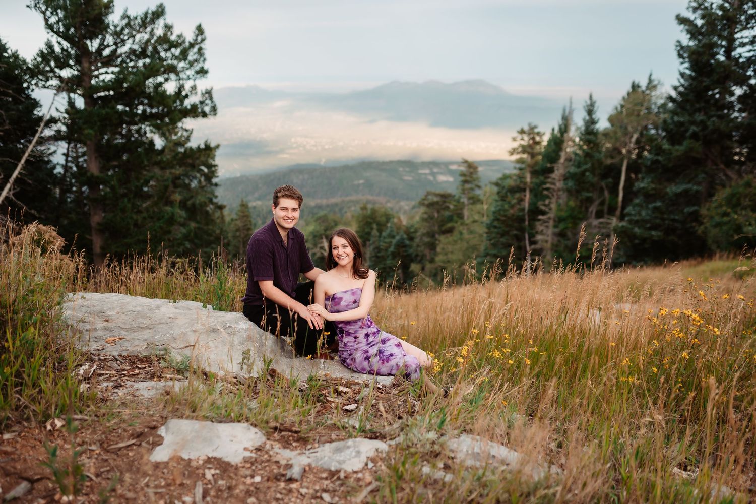 A couple sits together on rocky terrain with mountains and evergreen trees in the background.