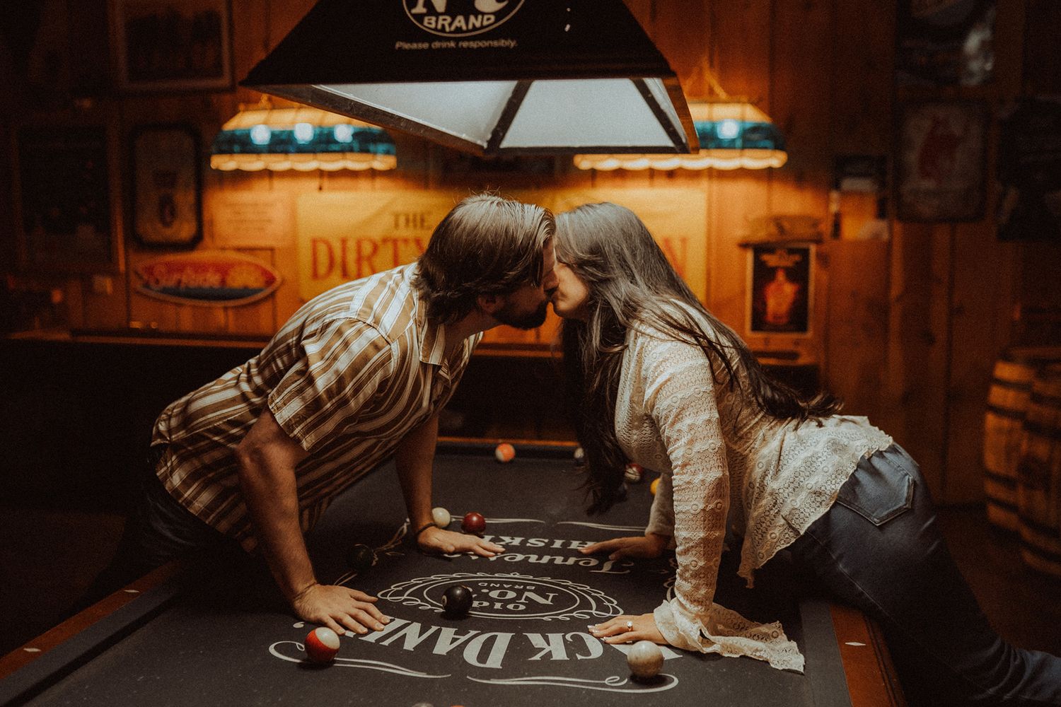 Couple sharing a romantic kiss leaning over a pool table in a cozy, warmly lit game room.