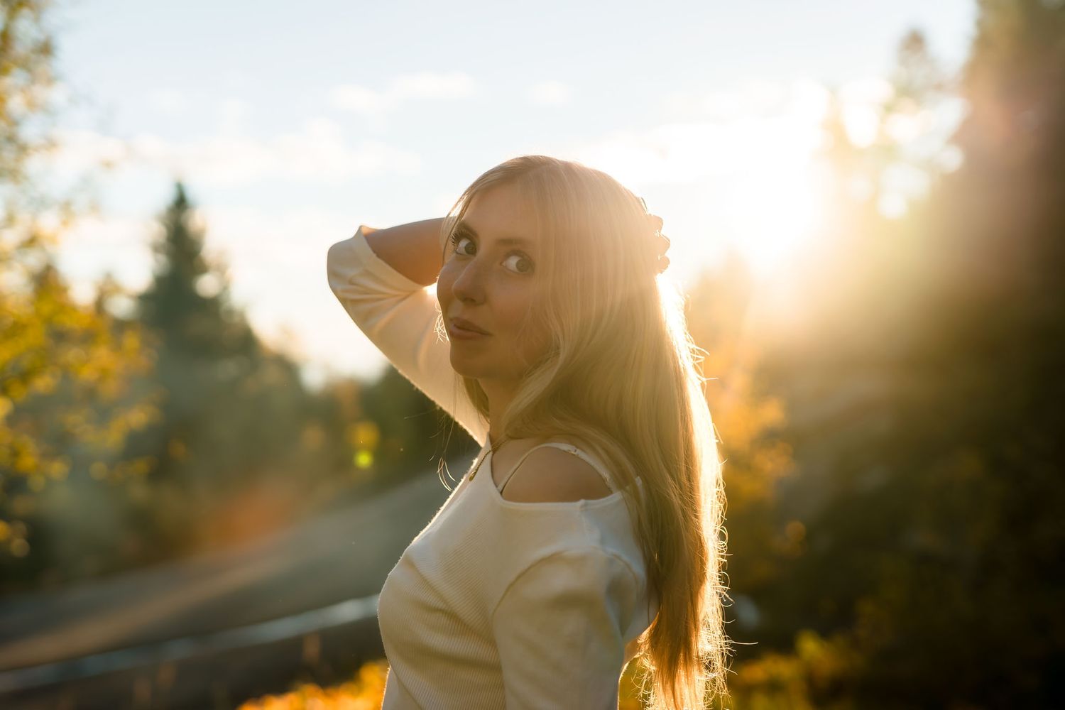 Young woman with long hair poses outdoors in golden sunset light, surrounded by autumn trees.