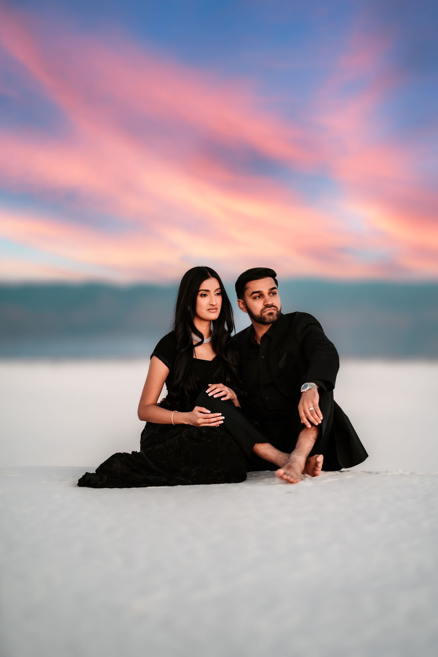A couple in black outfits poses romantically on white sand during a dreamy pink and blue sunset.
