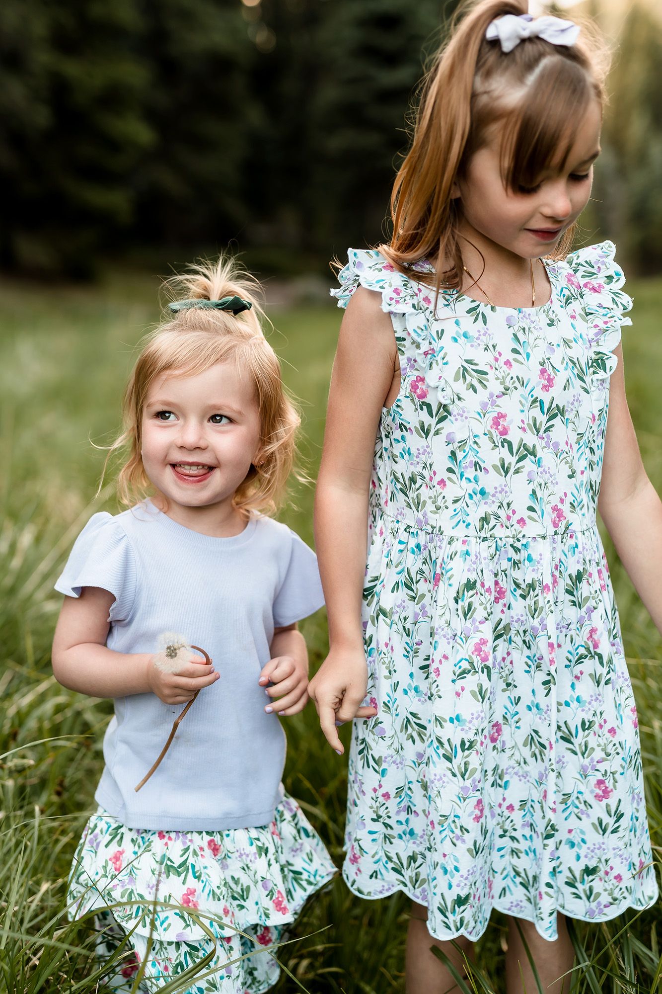 Two young girls in floral and blue outfits hold hands while playing outdoors in a grassy field.