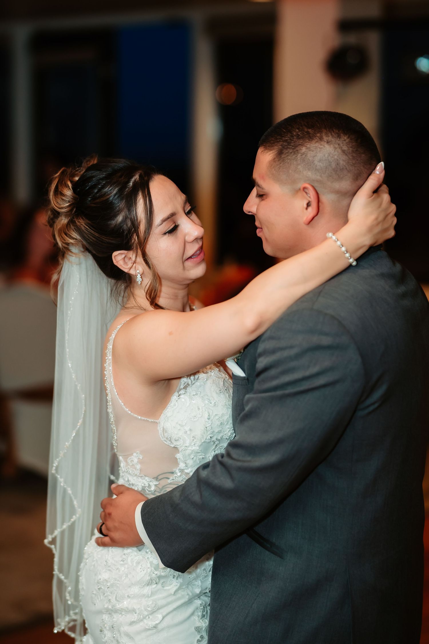 Bride in white lace gown dances intimately with groom in grey suit during their wedding reception.