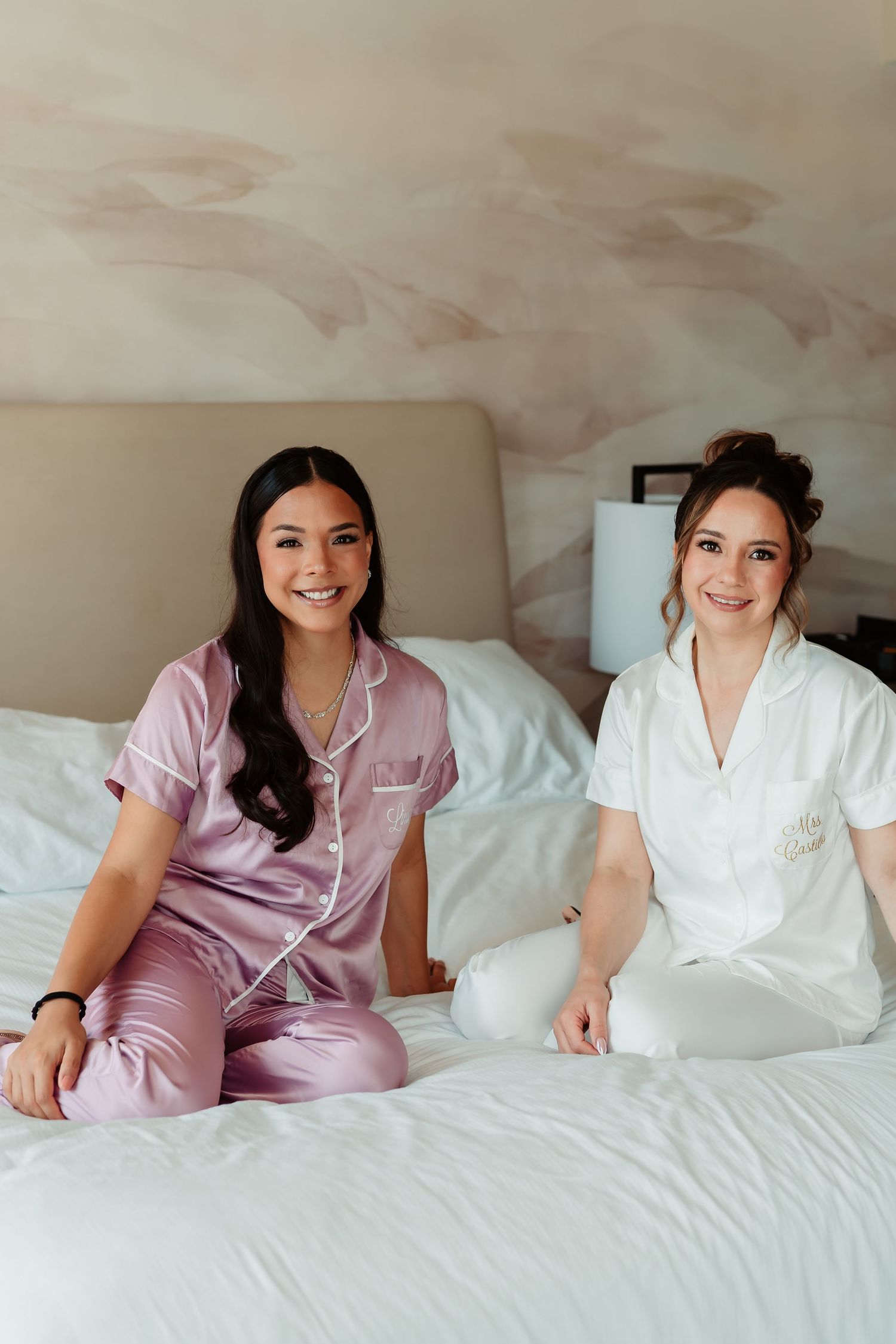 Two women wearing pink and white satin pajamas relaxing on a white bed during a slumber party.