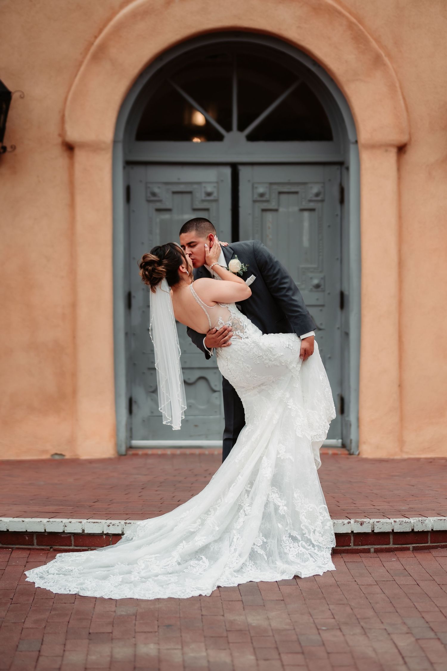 Elegant dance dips during wedding photoshoot against peach-colored church facade with grey doors.