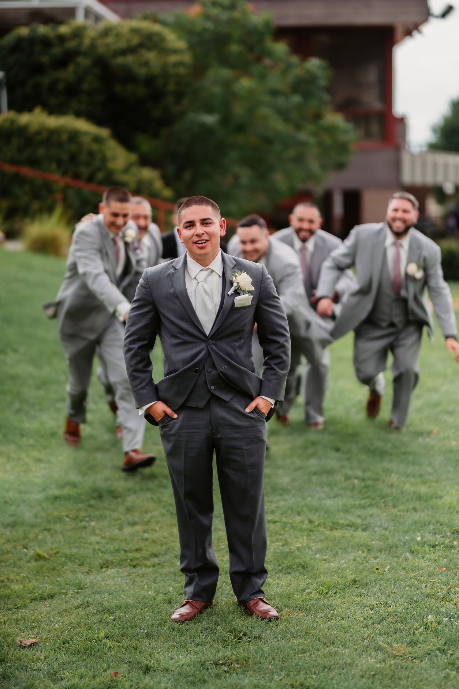 Groomsmen in gray suits run playfully across a grass lawn during wedding photos.