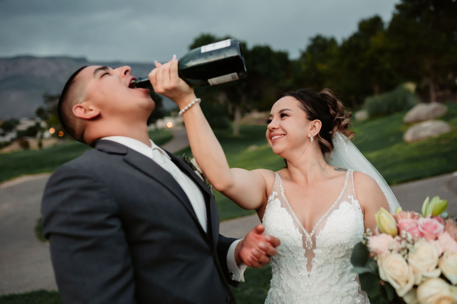A bride and groom share drinks while laughing together on their wedding day with mountains visible behind them.