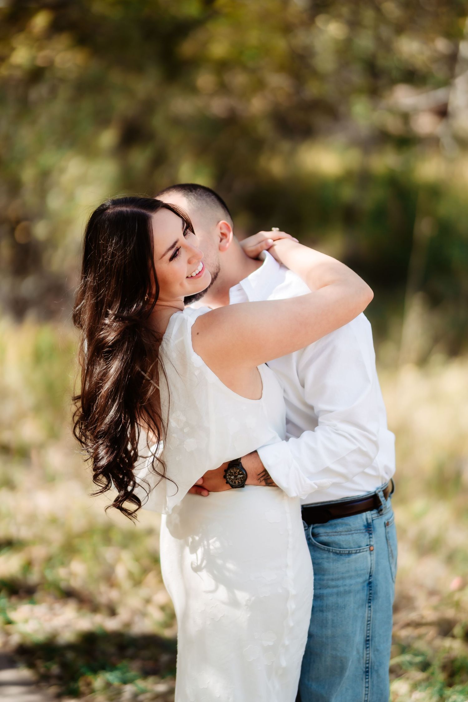 A couple in casual attire embraces outdoors in a natural setting with soft sunlight filtering through trees.