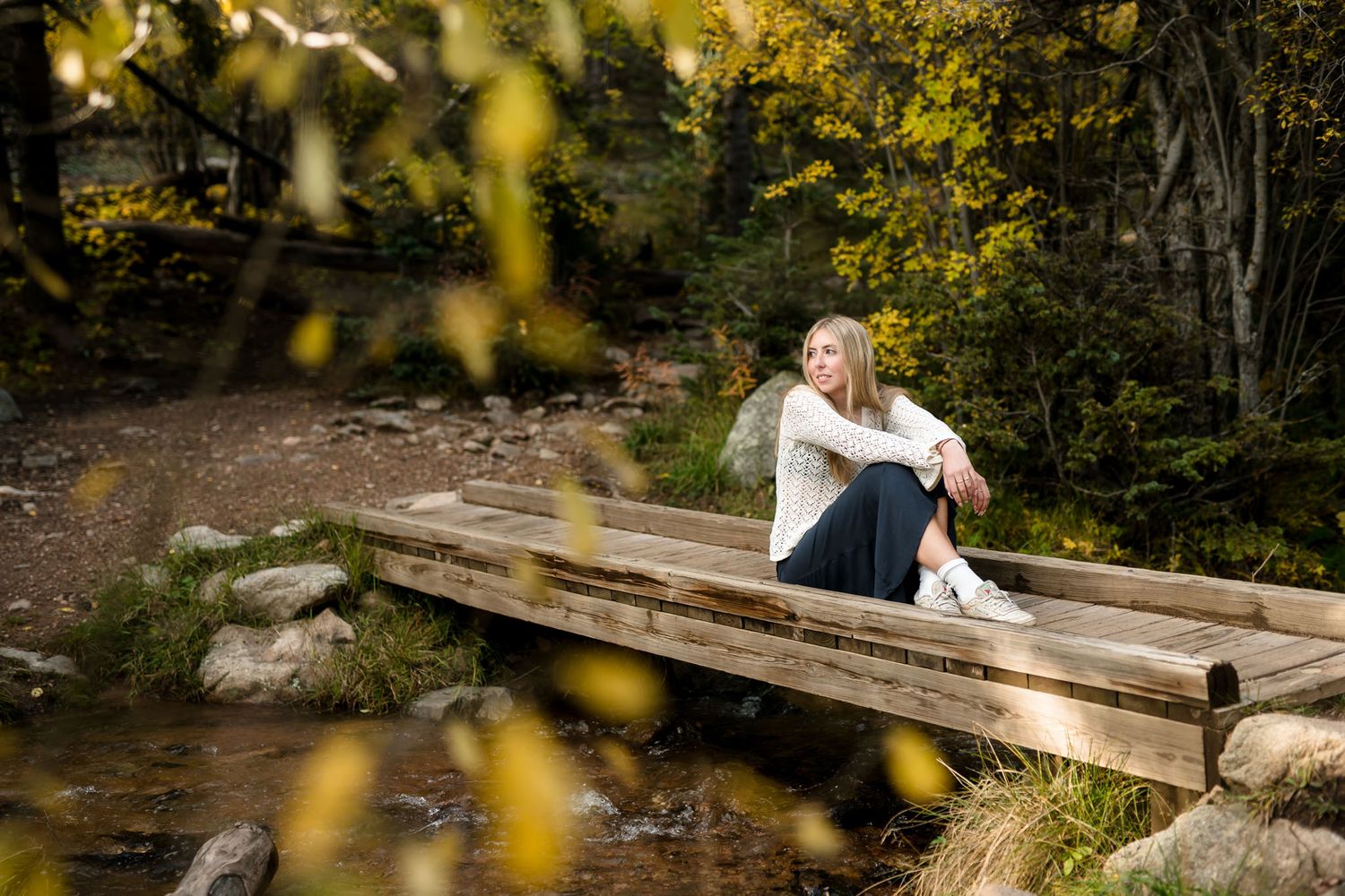 Young blonde woman sitting on a wooden bridge surrounded by autumn foliage in a serene forest setting.