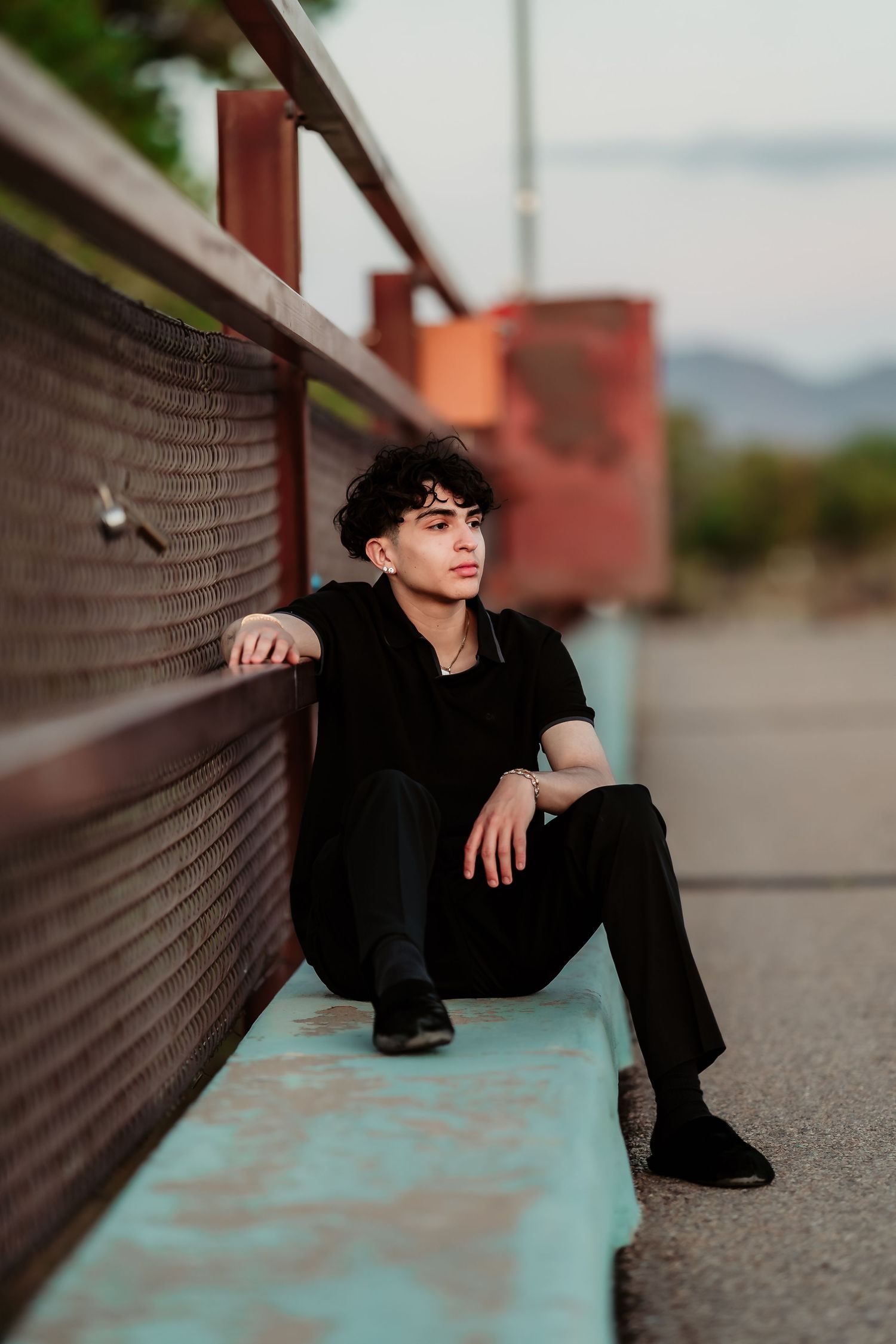 Person in all black outfit sitting on turquoise ledge against industrial background at dusk.