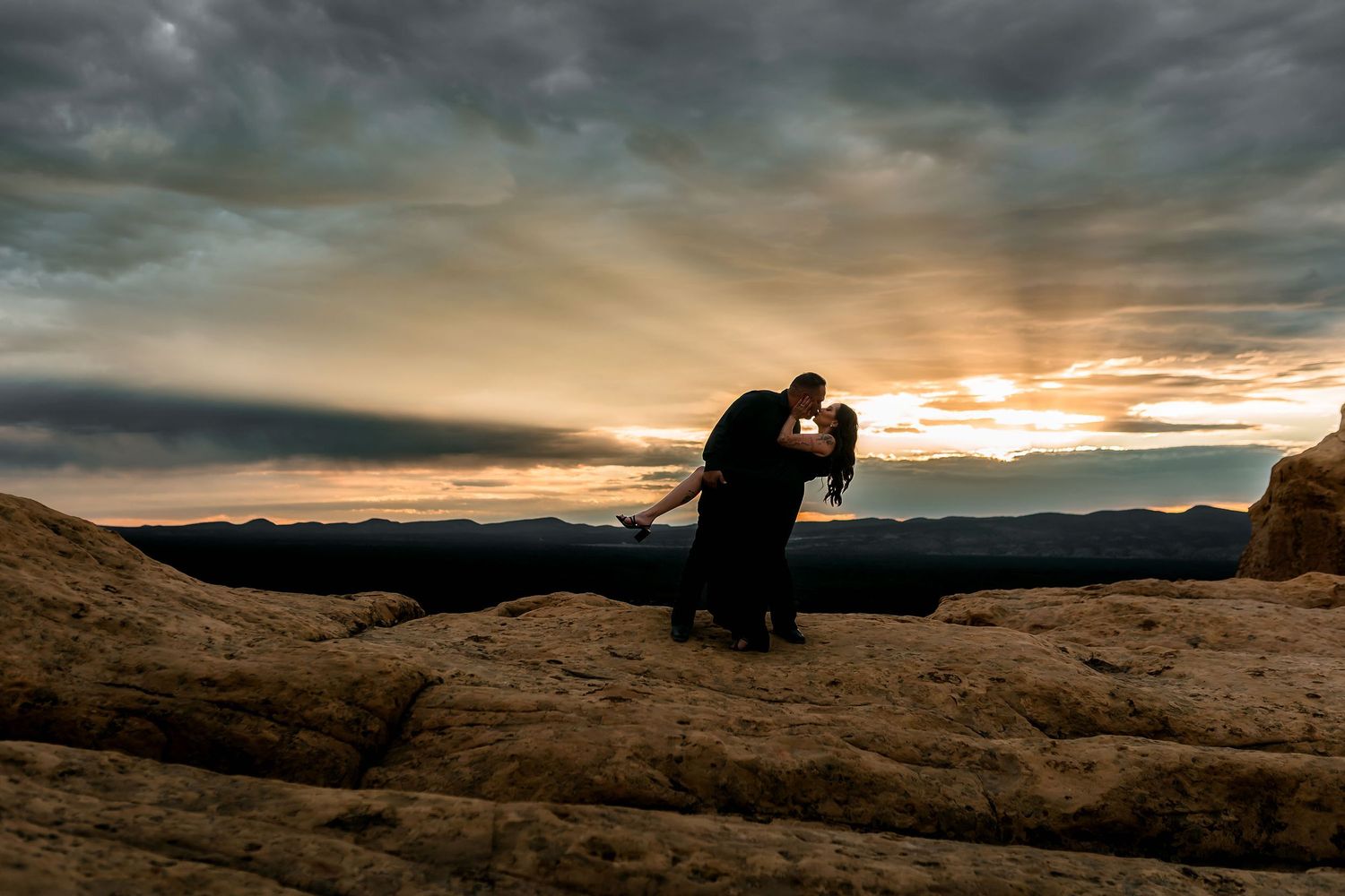 Couple sharing a romantic kiss silhouetted against a stunning desert sunset on rocky terrain.