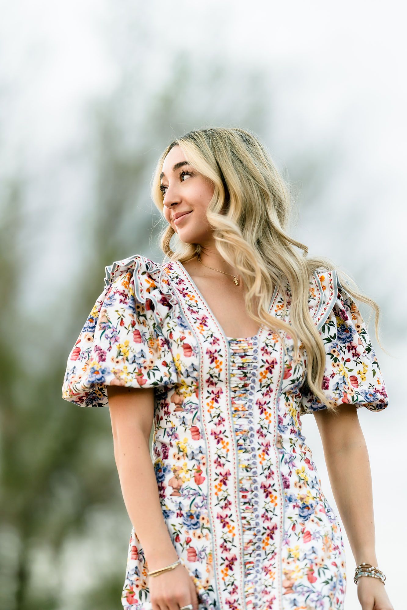Young woman in floral puff-sleeve dress smiling outdoors with blurred green trees in background.