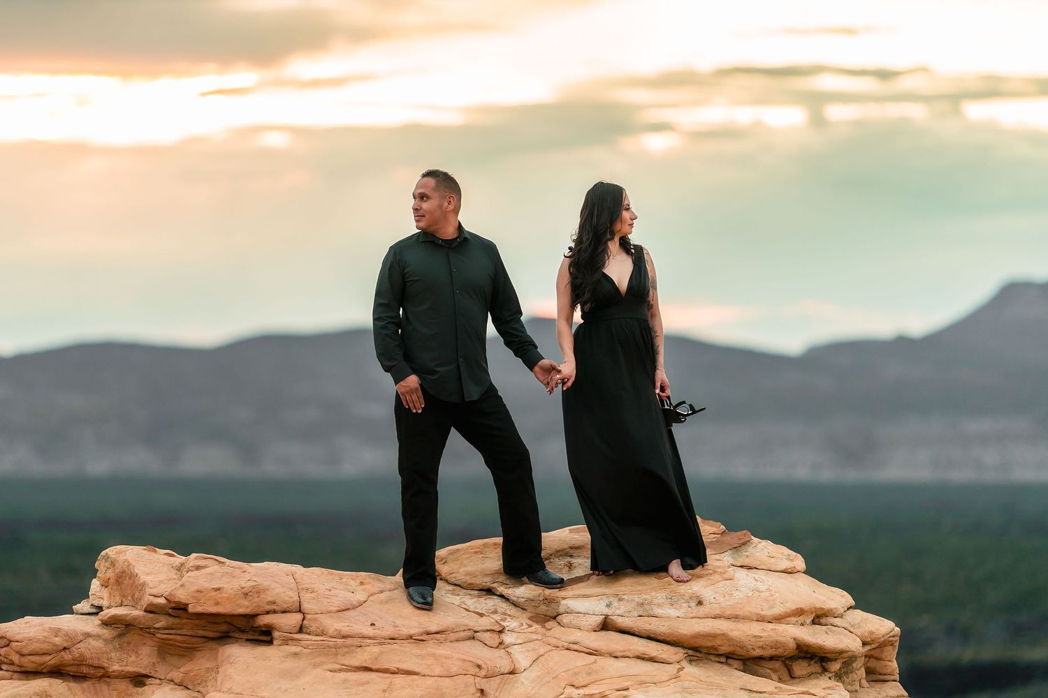Couple holding hands on desert rock formation at golden hour with scenic mountain backdrop.