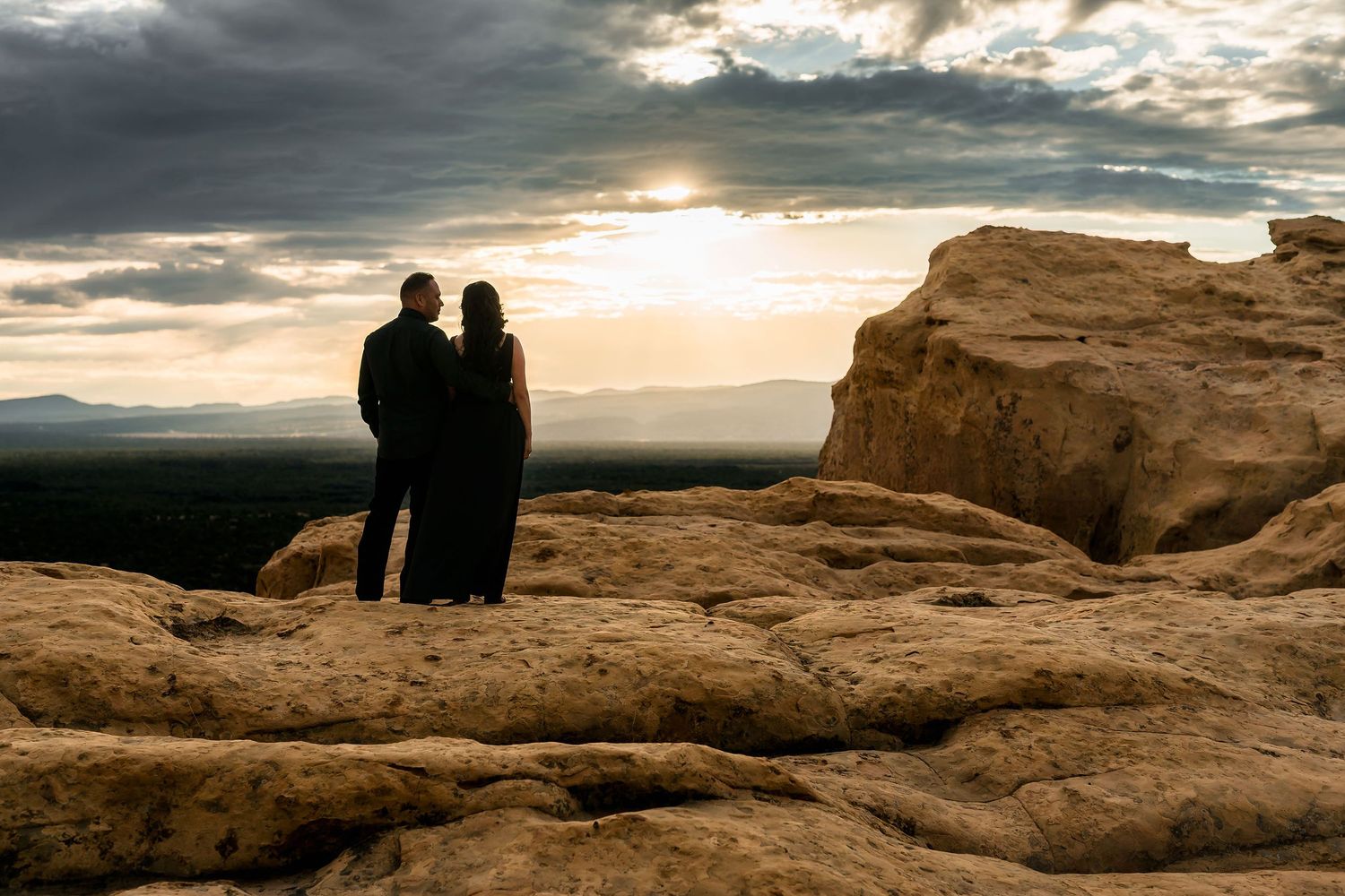 Couple standing together on sandstone rocks watching a dramatic golden sunset over a sweeping desert vista.