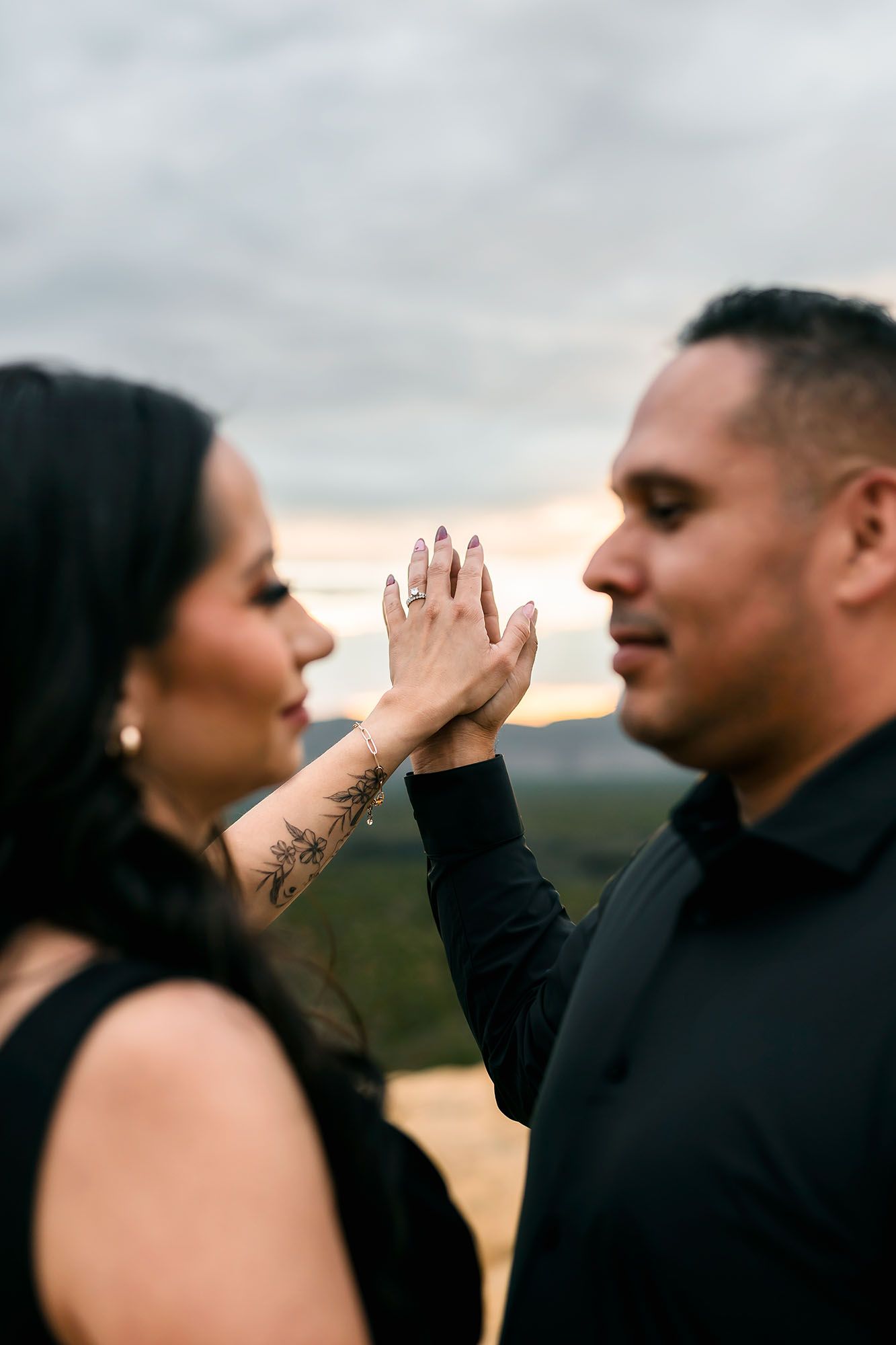 Couple facing each other at golden hour, woman raising tattooed hand showing engagement ring.