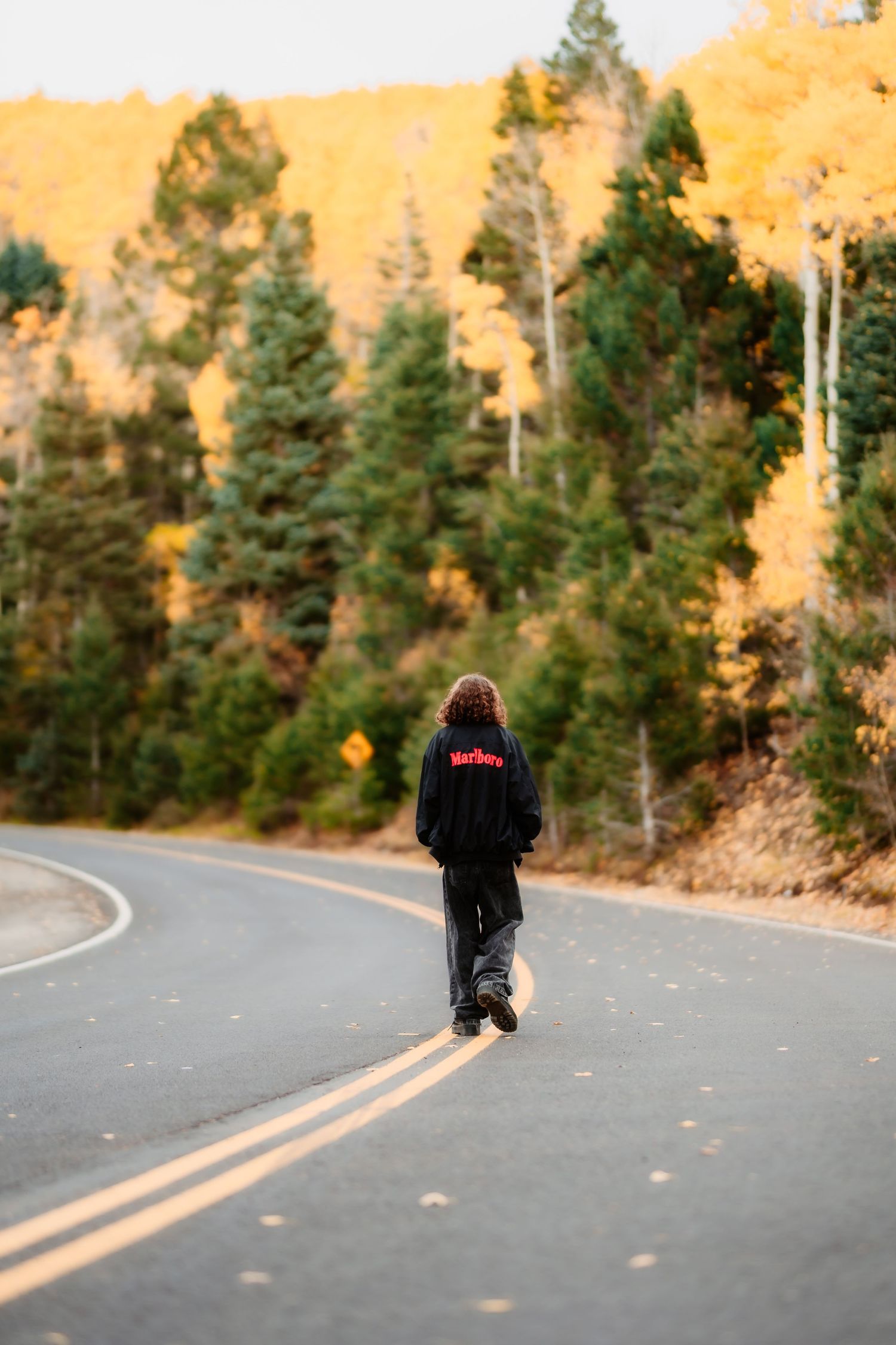 A person in dark clothing walks down a winding mountain road with autumn trees and golden hills in the background.