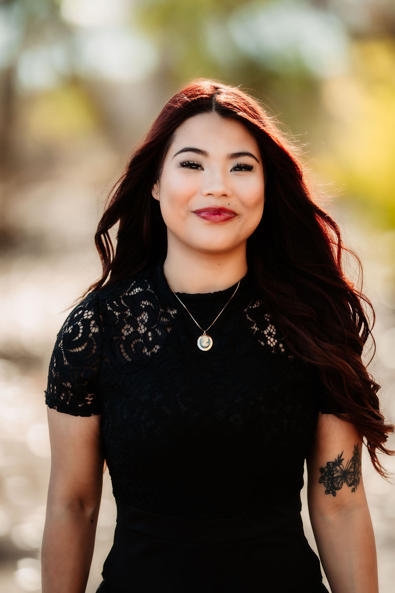 Woman with flowing dark hair wearing black lace top poses against blurred outdoor background.