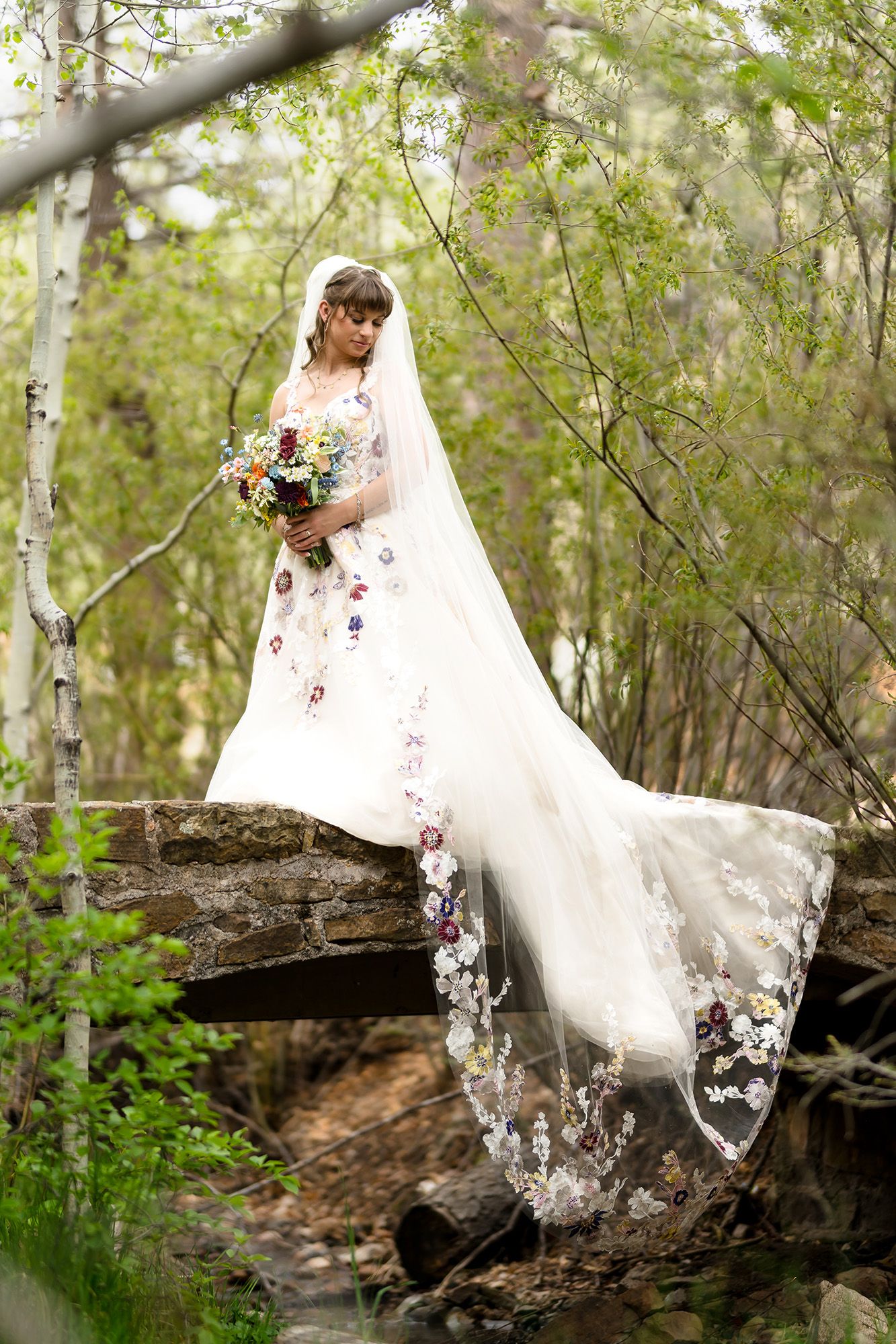 Bride in floral embroidered wedding gown and veil holds bouquet on stone bridge surrounded by lush green forest trees.