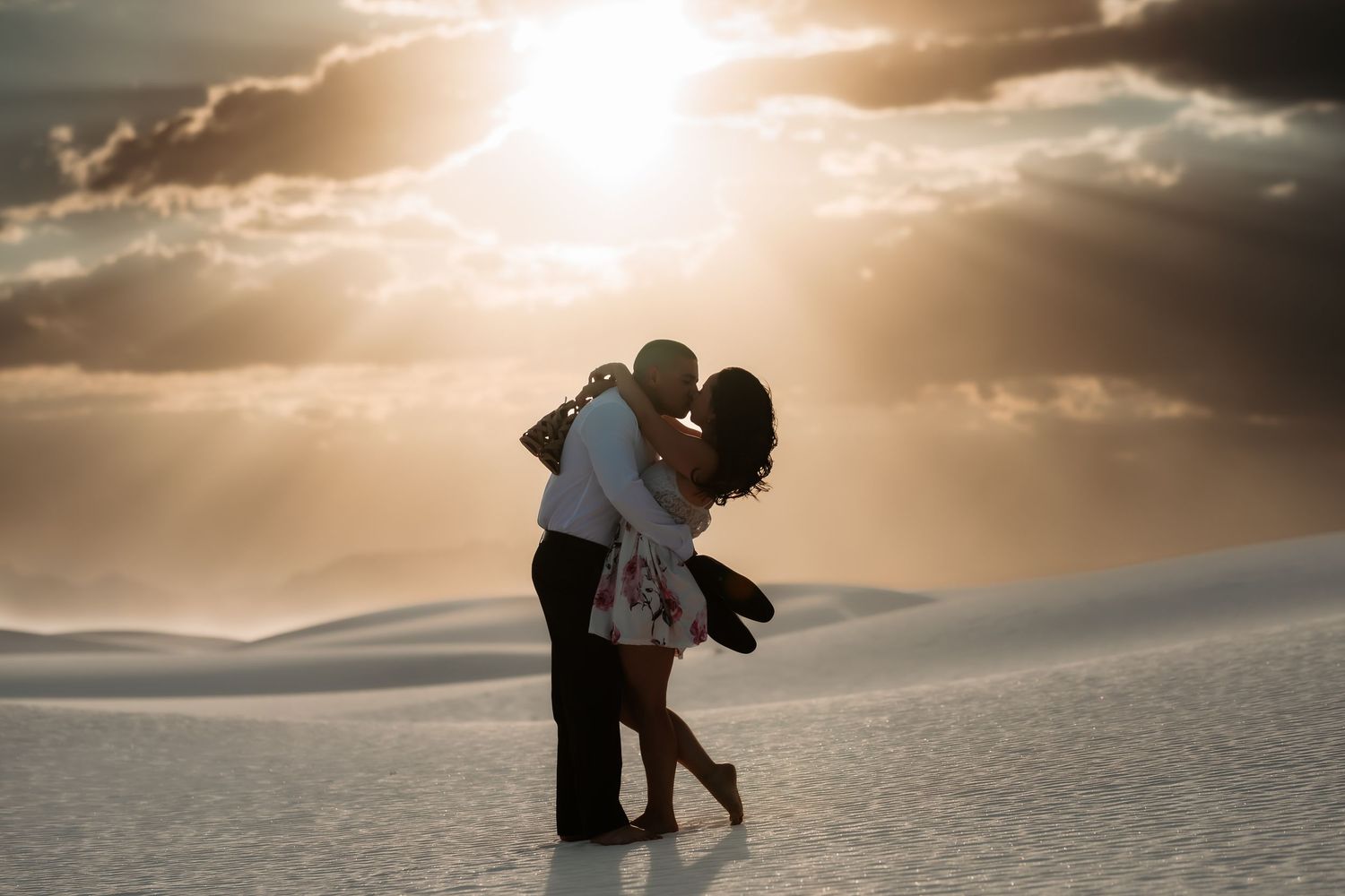 Two silhouettes embrace against a dramatic sunset sky at White Sands National Park with sunbeams breaking through clouds.