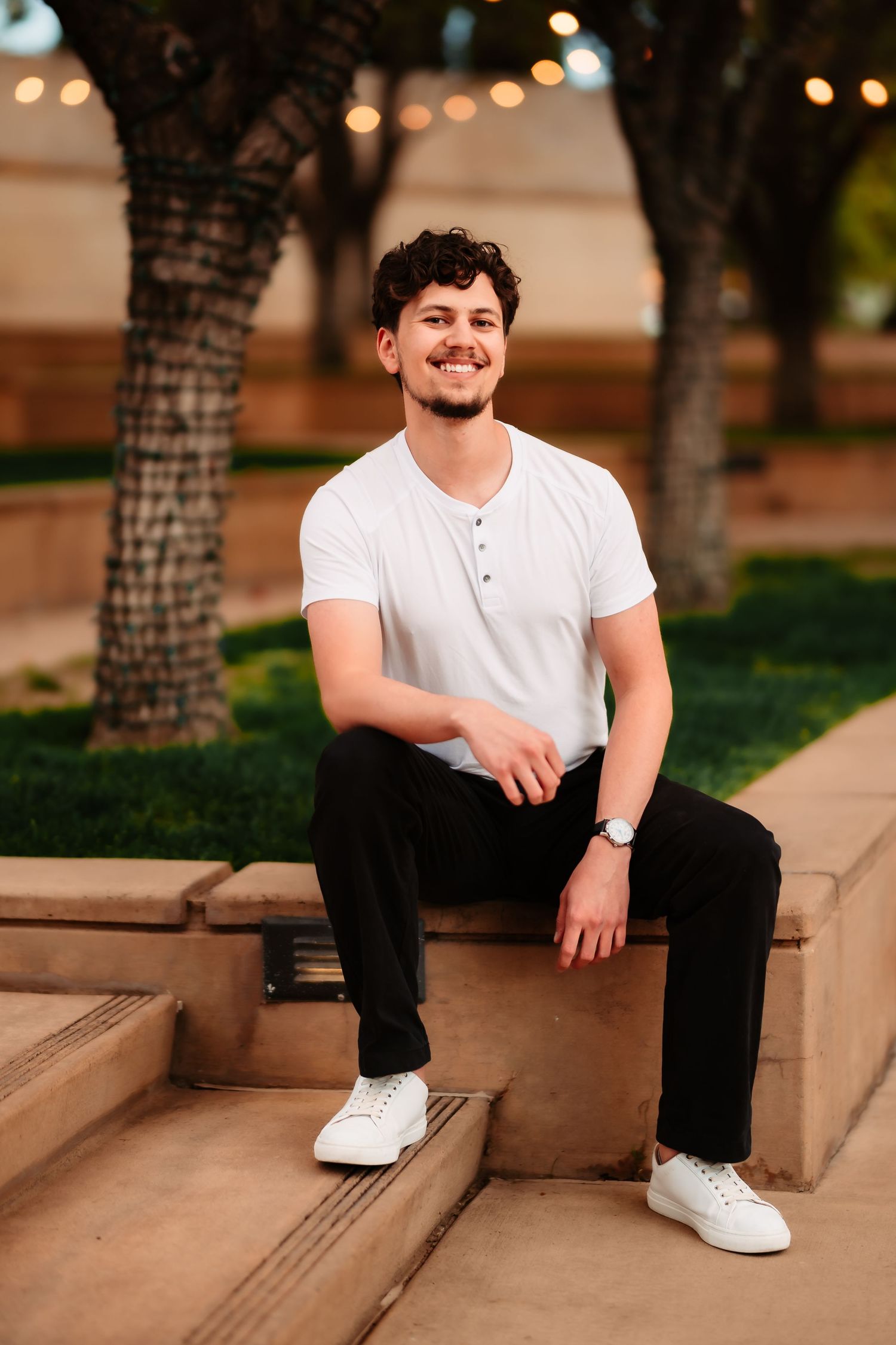 A person in a white t-shirt and black pants sits casually on concrete steps outdoors with string lights in the background.