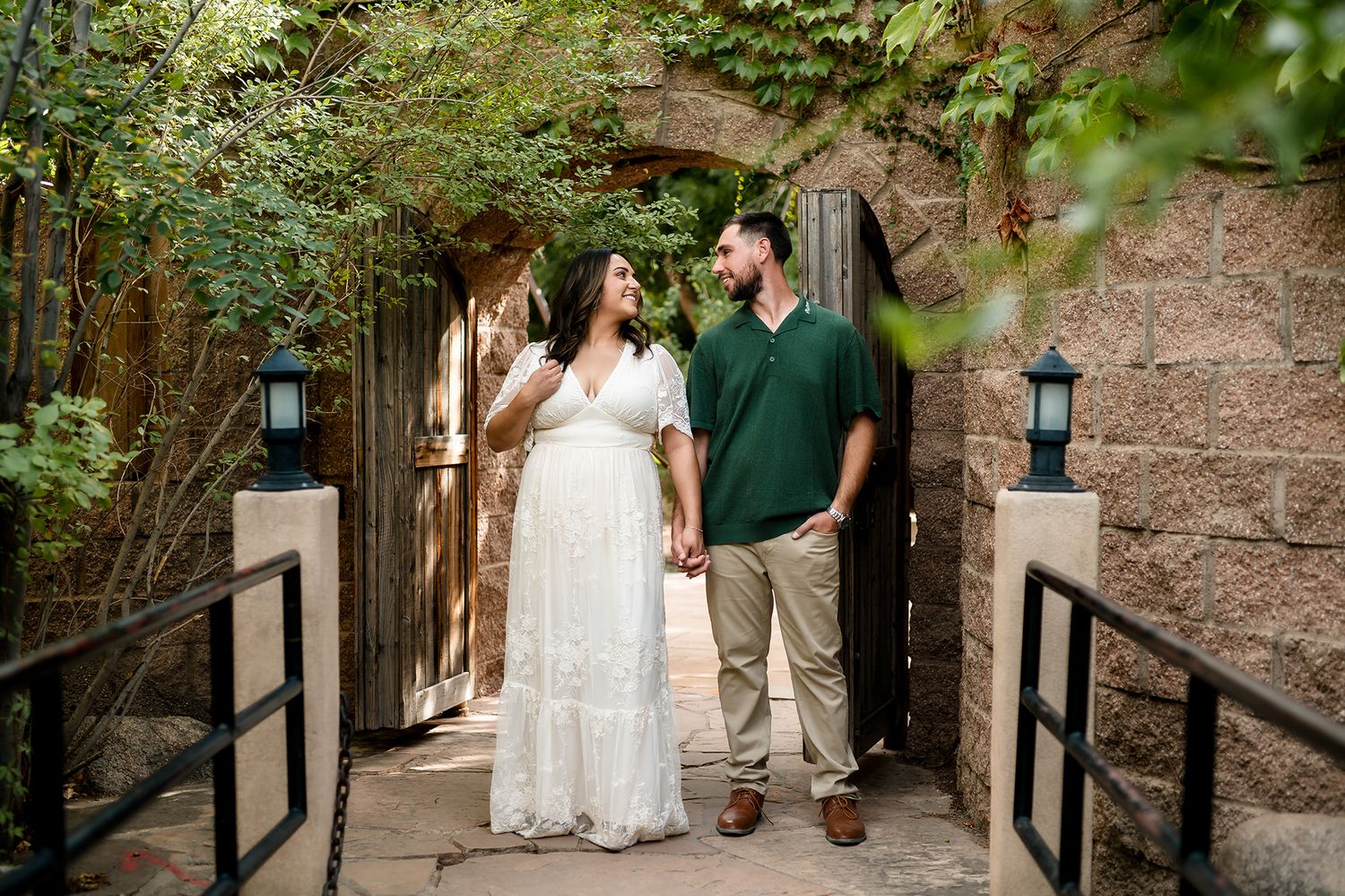 Couple holding hands and smiling at each other under a stone archway covered in ivy.