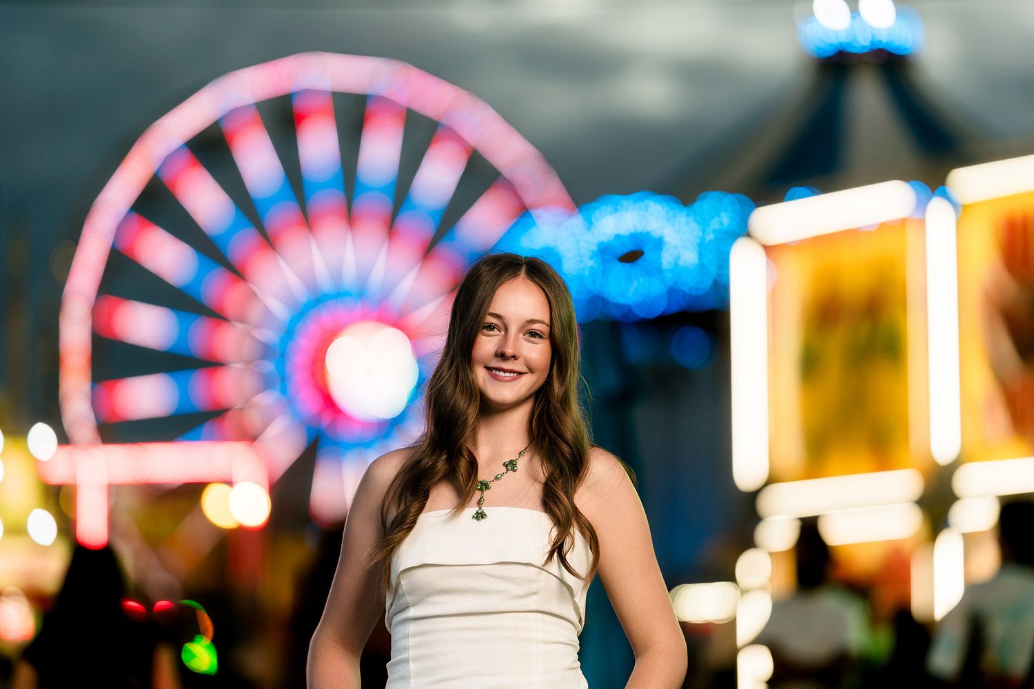 Young woman in white dress smiling at a carnival with a lit Ferris wheel and colorful lights at night.