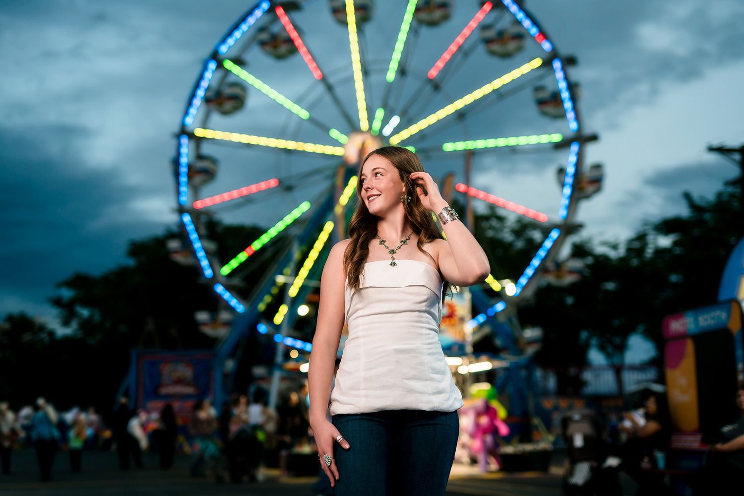 Woman standing in front of a colorful illuminated Ferris wheel at a fairground during dusk.