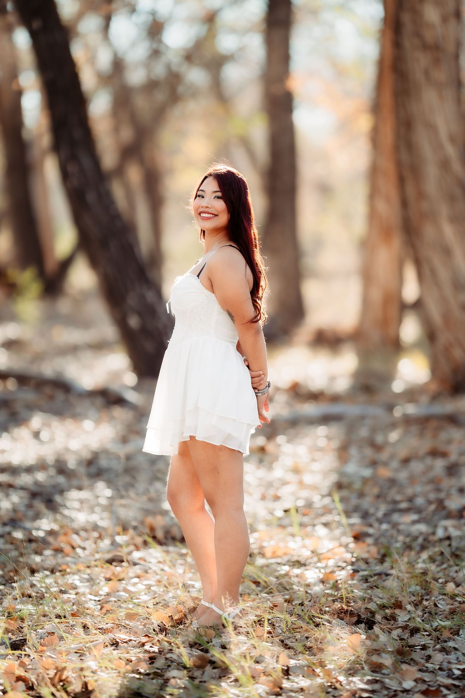 Woman in white dress poses gracefully in autumn forest with fallen leaves and soft natural lighting.