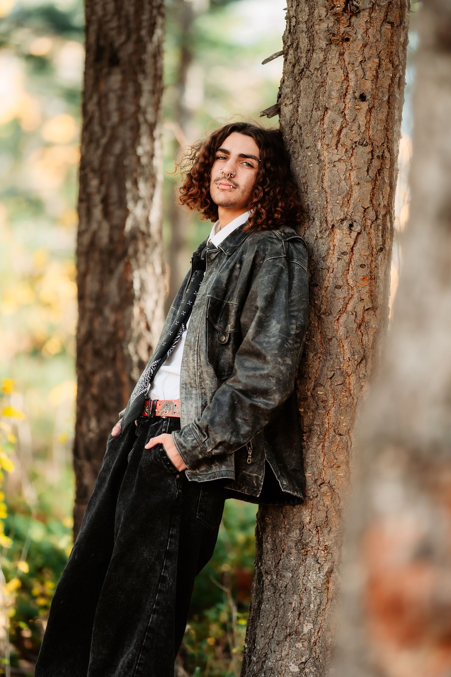 Person with curly hair wearing a black leather jacket poses against a tree trunk in an autumn forest setting.
